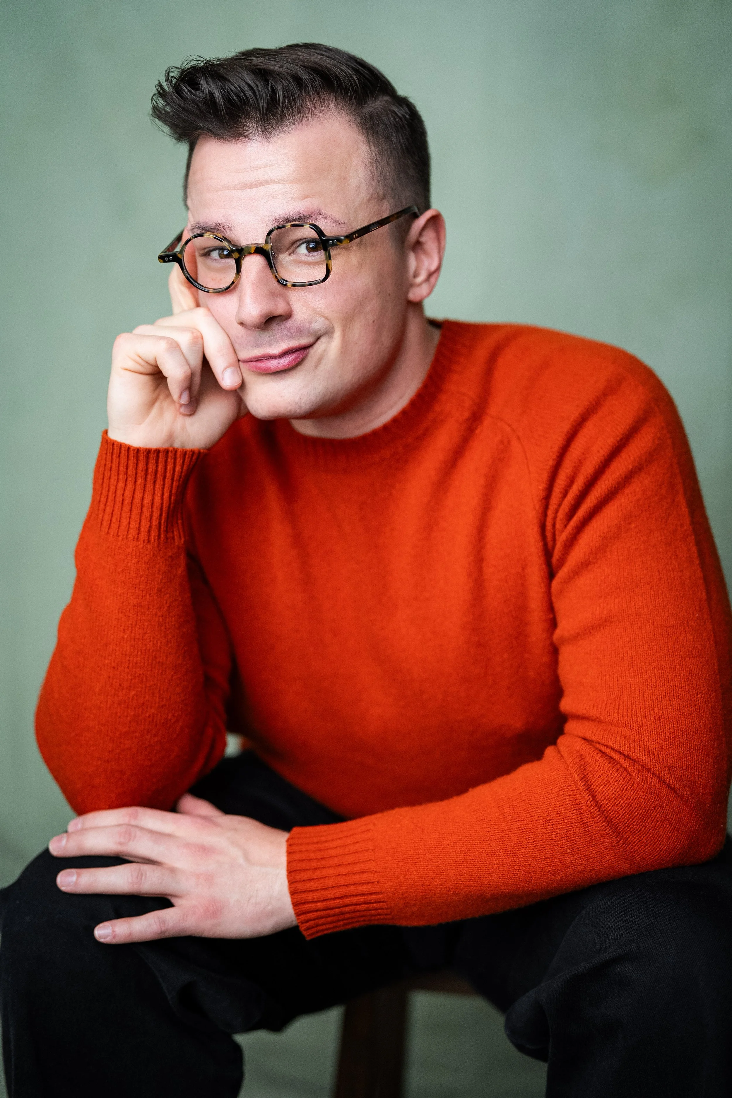 A headshot of RP McLaughlin. They are wearing a burnt orange sweater and black pants. They are resting their he'd in their right hand.  Credit: YellowBelly