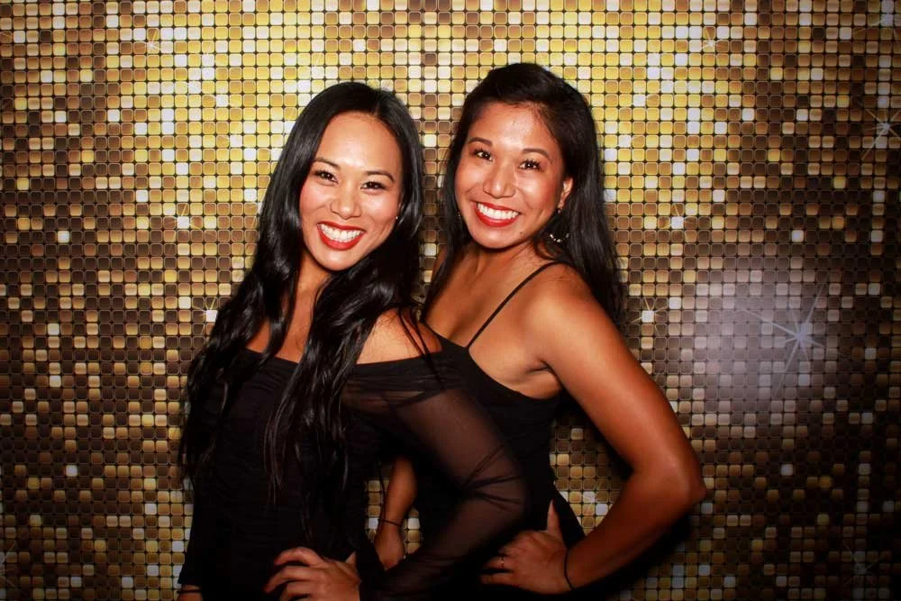 Guests posing at a Cheers To You open-air photo booth with a gold sequin backdrop