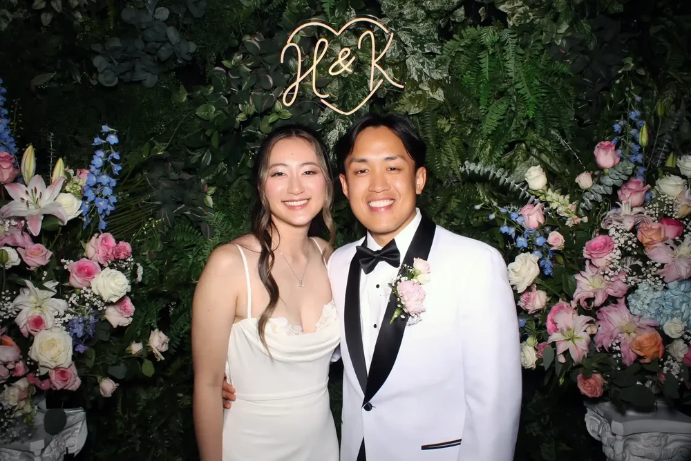 Couple at a Cheers To You wedding photo booth with a floral backdrop and neon monogram