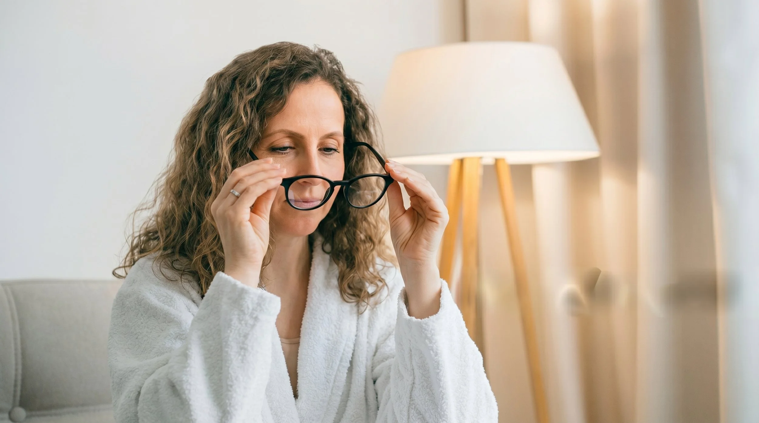 Woman with curly hair and glasses adjusting them while sitting on a couch with a lamp in the background.