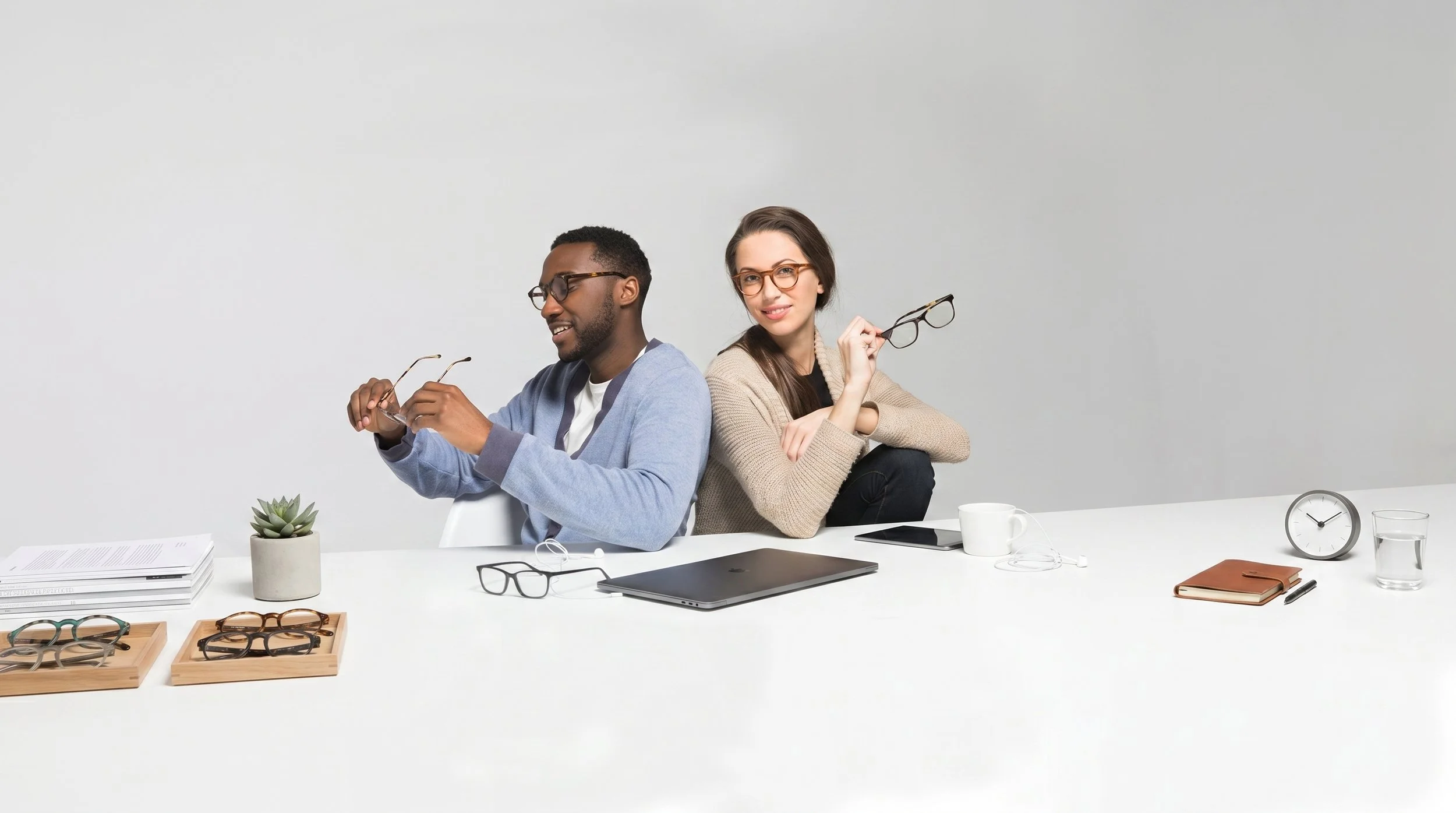 Two people, a man and a woman, sit back-to-back at a white desk with glasses, a laptop, notebooks, and office supplies, against a plain light gray background.