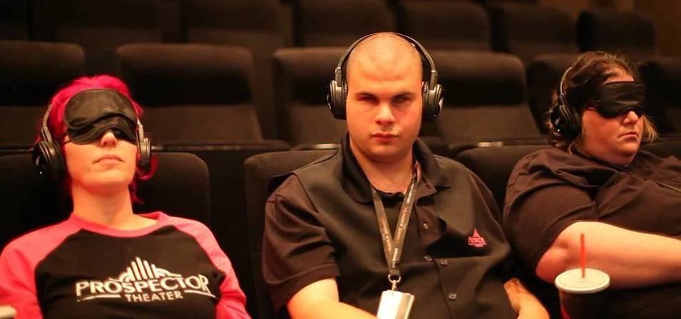Two women sit in theater seats wearing blindfolds. A man wearing headphones sits in between them.