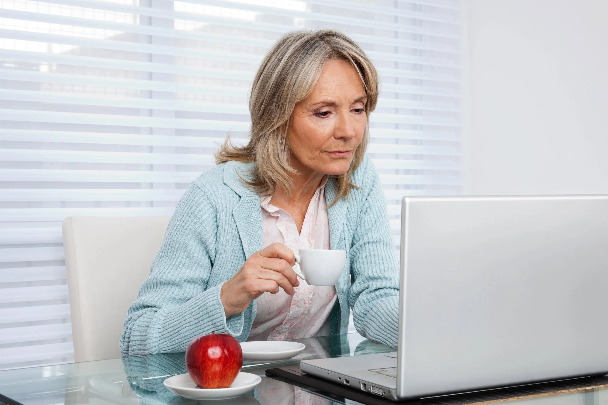 Older adult sitting at a table using a laptop while holding a cup of coffee, with an apple and plate in the foreground.