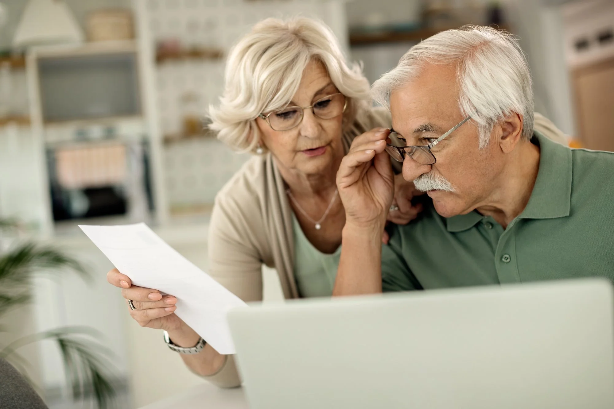 Older couple reviewing financial documents together at home, focusing on retirement planning, insurance decisions, and household finances using a laptop and paperwork.