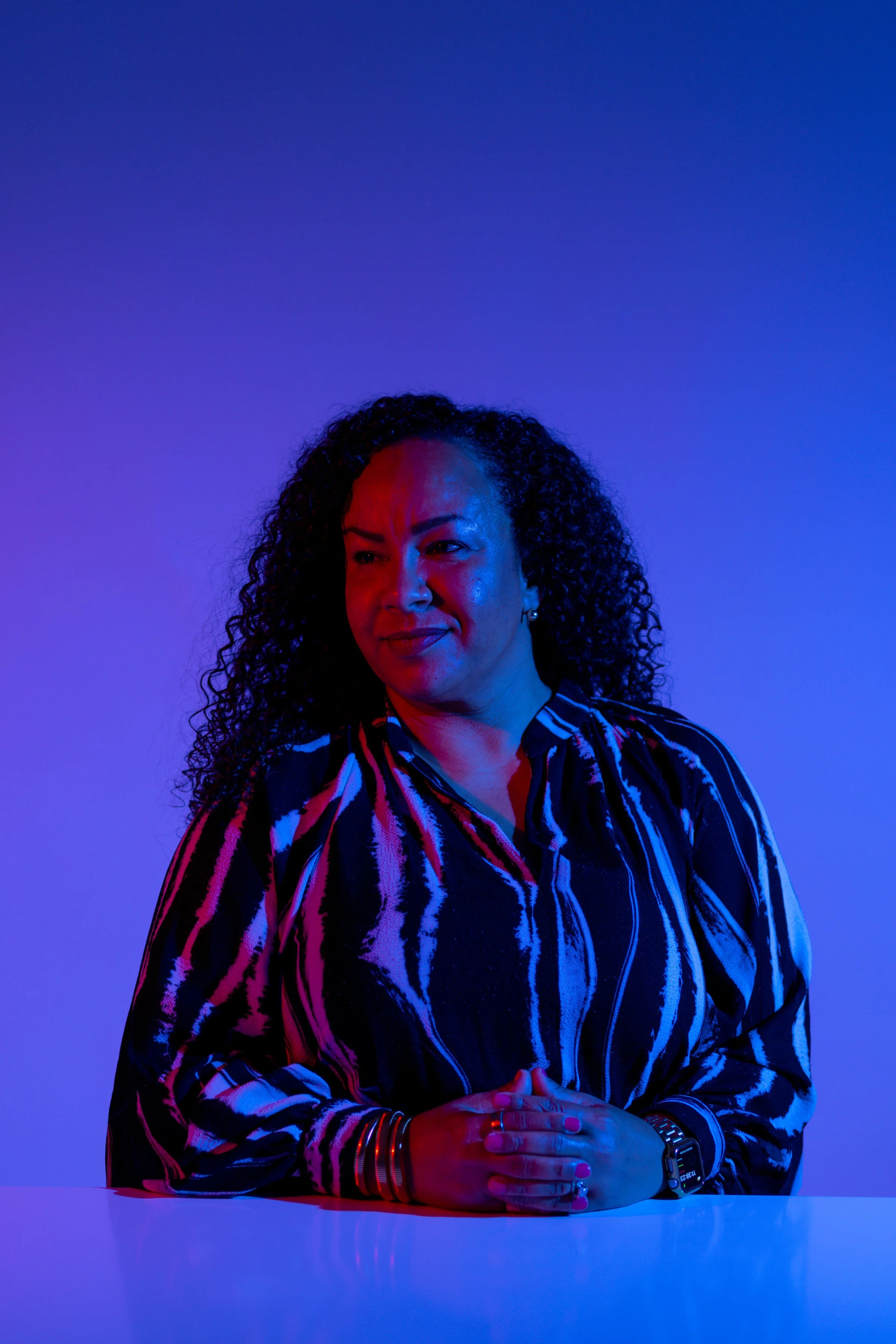 A woman with curly hair wearing a black and white striped shirt, sitting at a white table with her hands clasped, softly smiling, illuminated with red and blue lighting against a plain background.