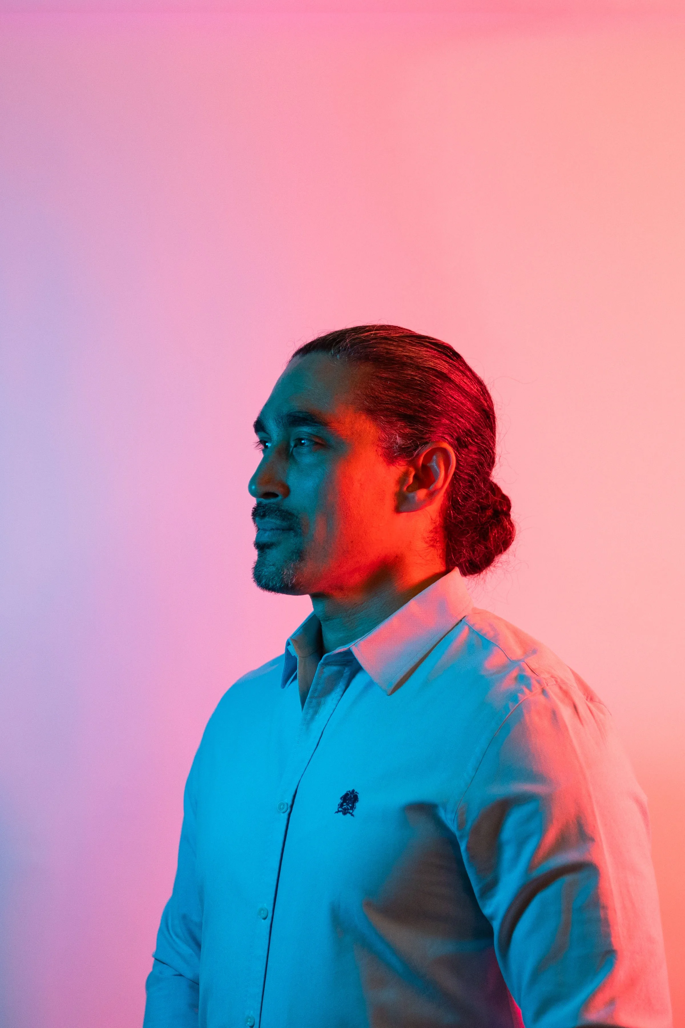 A man with tied-back dark hair and facial hair, wearing a light-colored button-down shirt, looking to the left, illuminated by colorful lighting in pink and blue hues.