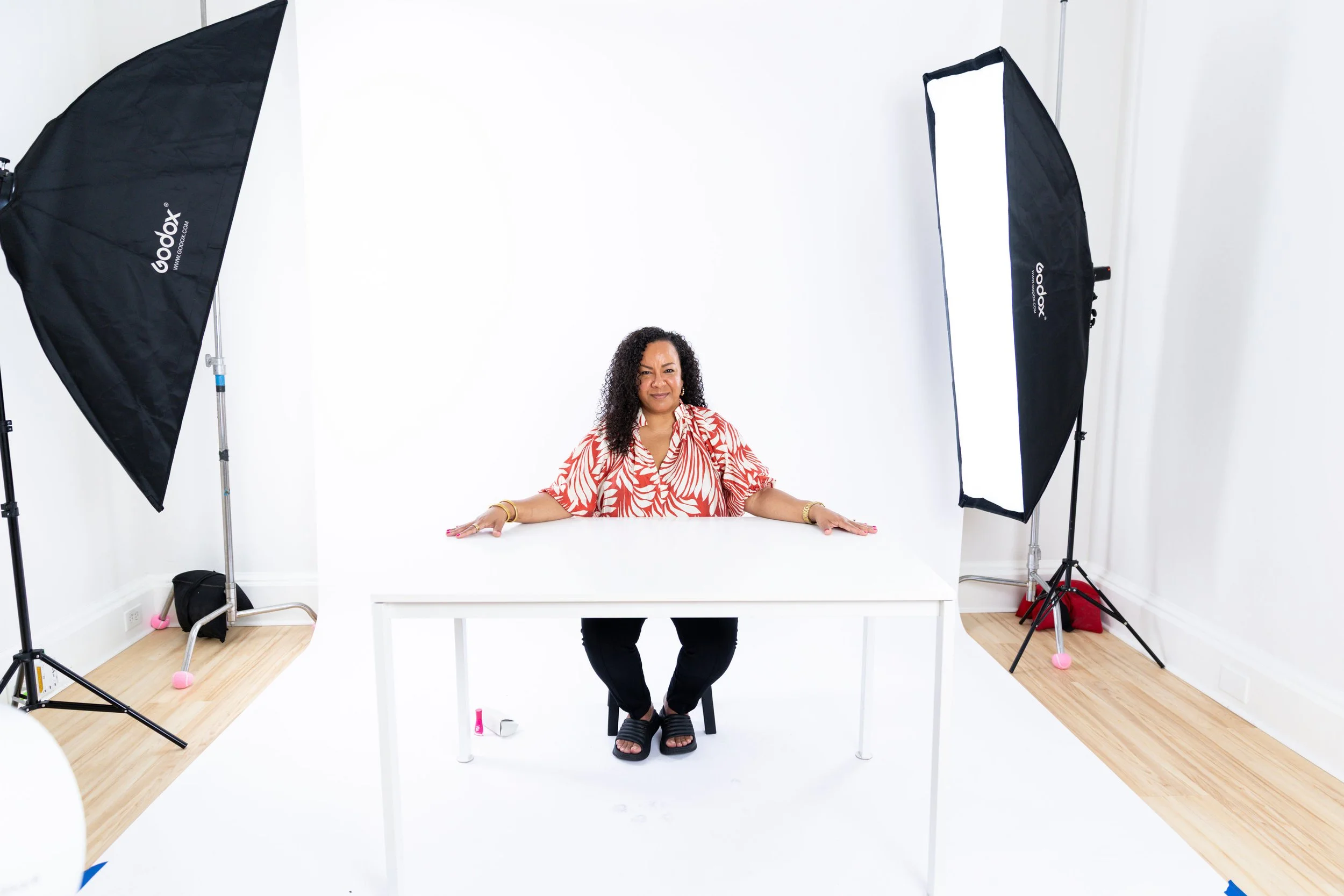 Woman sitting at a white table in a photography studio with a white background and professional lighting equipment.