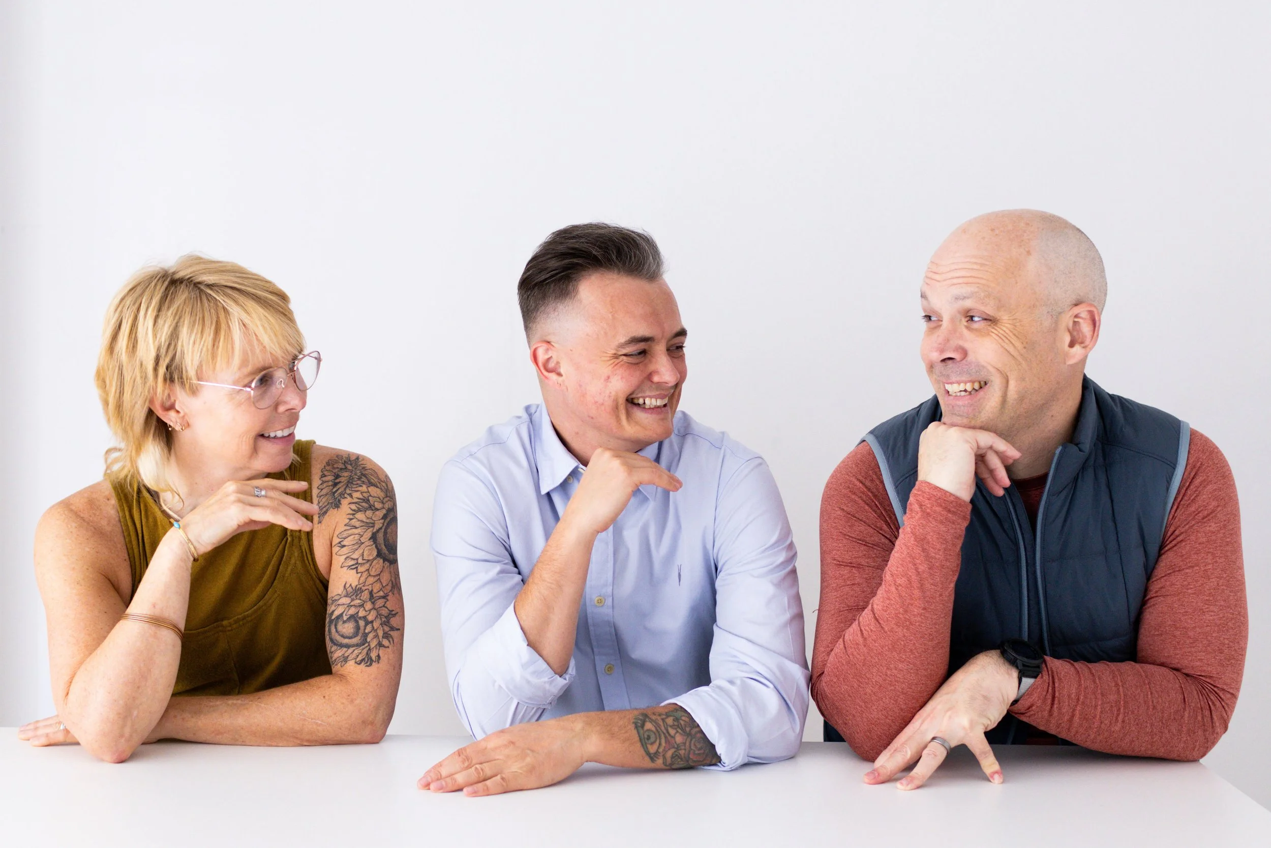 Three adults sitting at a table, smiling and talking to each other, against a white background.