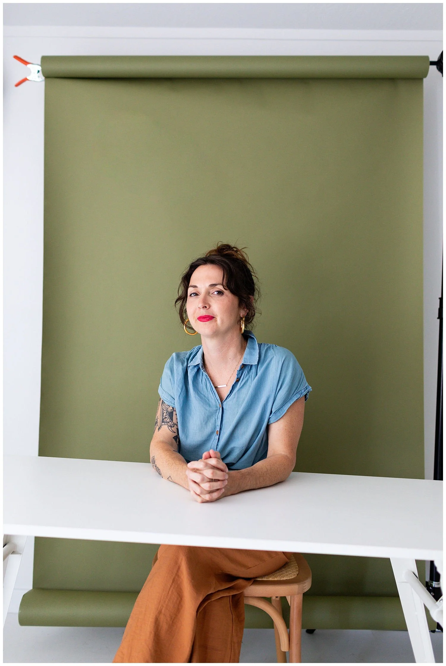 A woman with curly dark hair and tattoos on her left arm sits at a white table in front of a green backdrop, wearing a light blue shirt and brown pants.