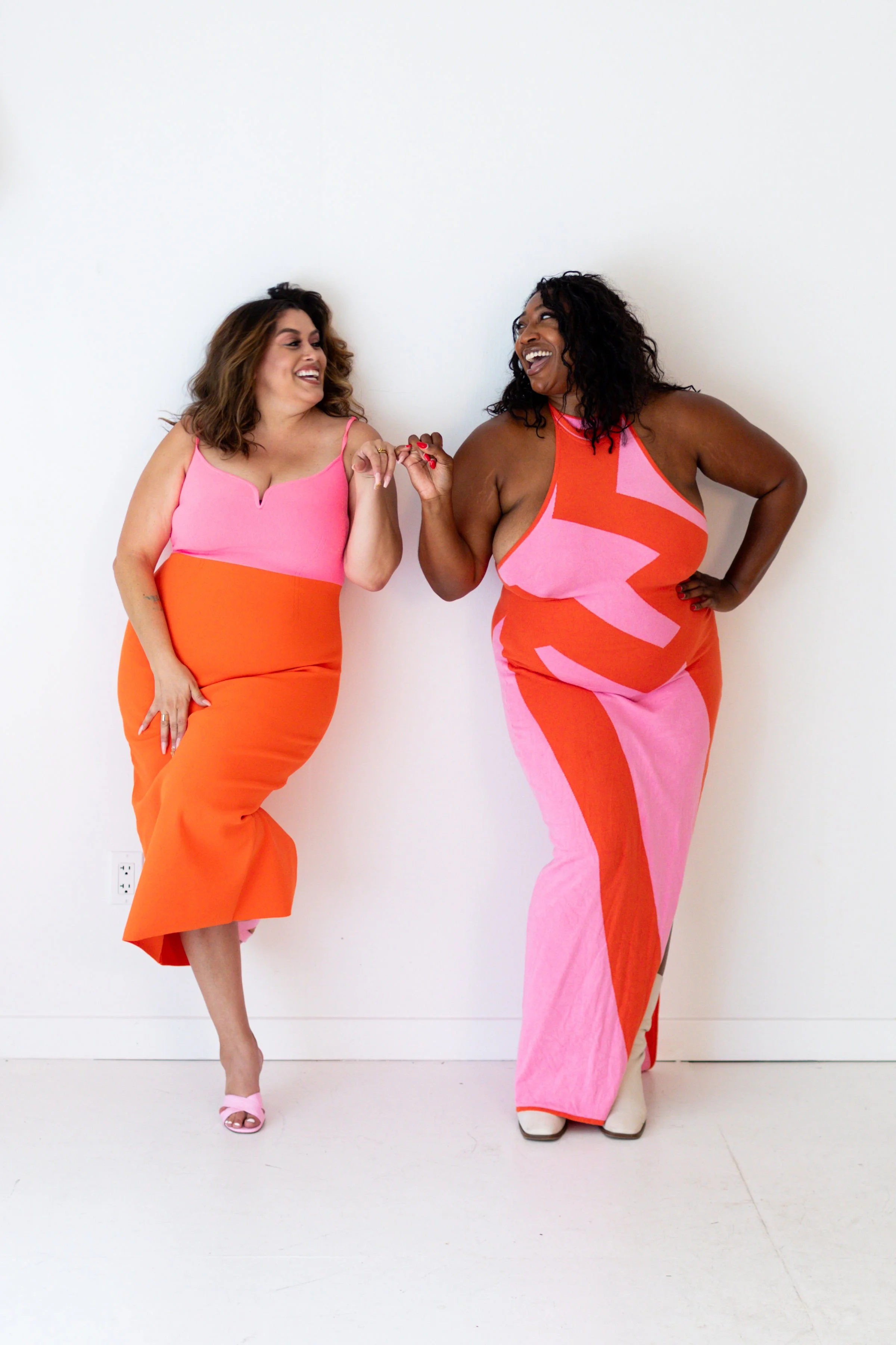 Two women wearing matching color-block dresses in pink and orange, standing against a white wall, smiling and holding hands.