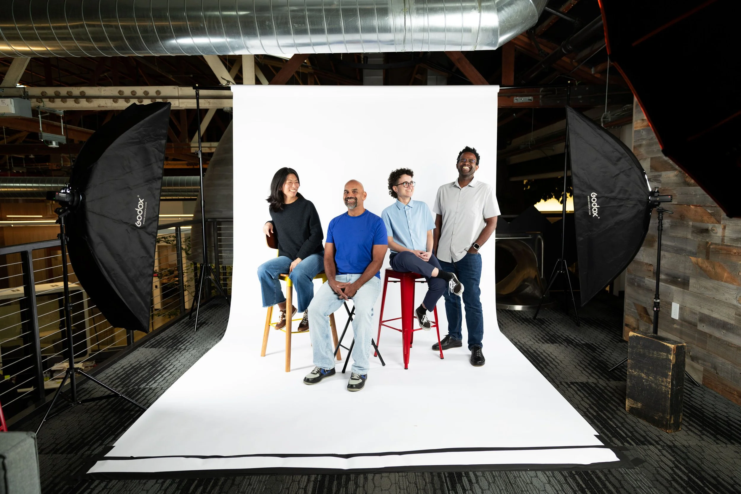 Four people, two women and two men, sitting and standing on stools in a photo studio with white backdrop, surrounded by studio lighting equipment.