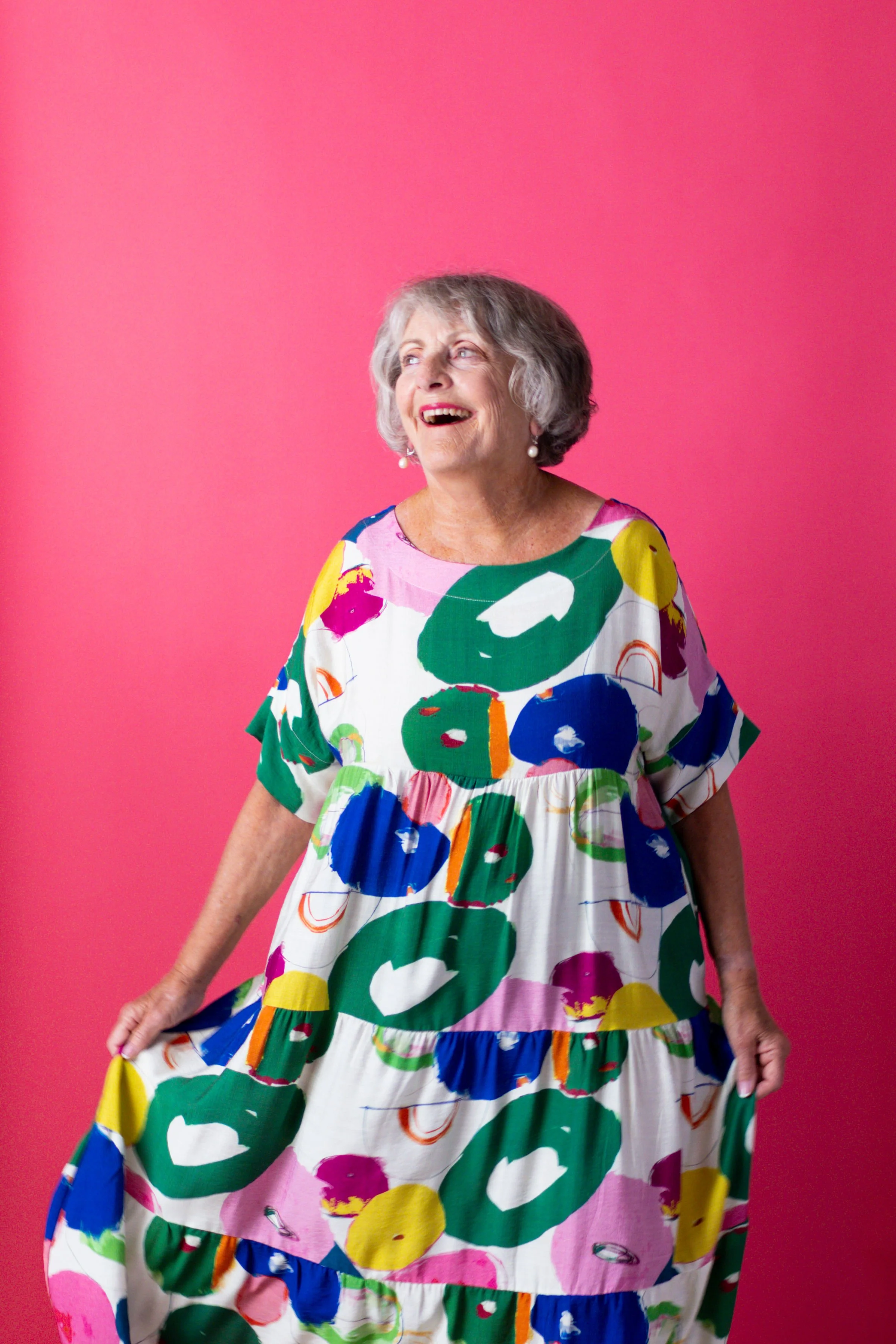 An elderly woman with gray hair wearing a colorful, patterned dress standing against a pink background, smiling and looking off to the side.