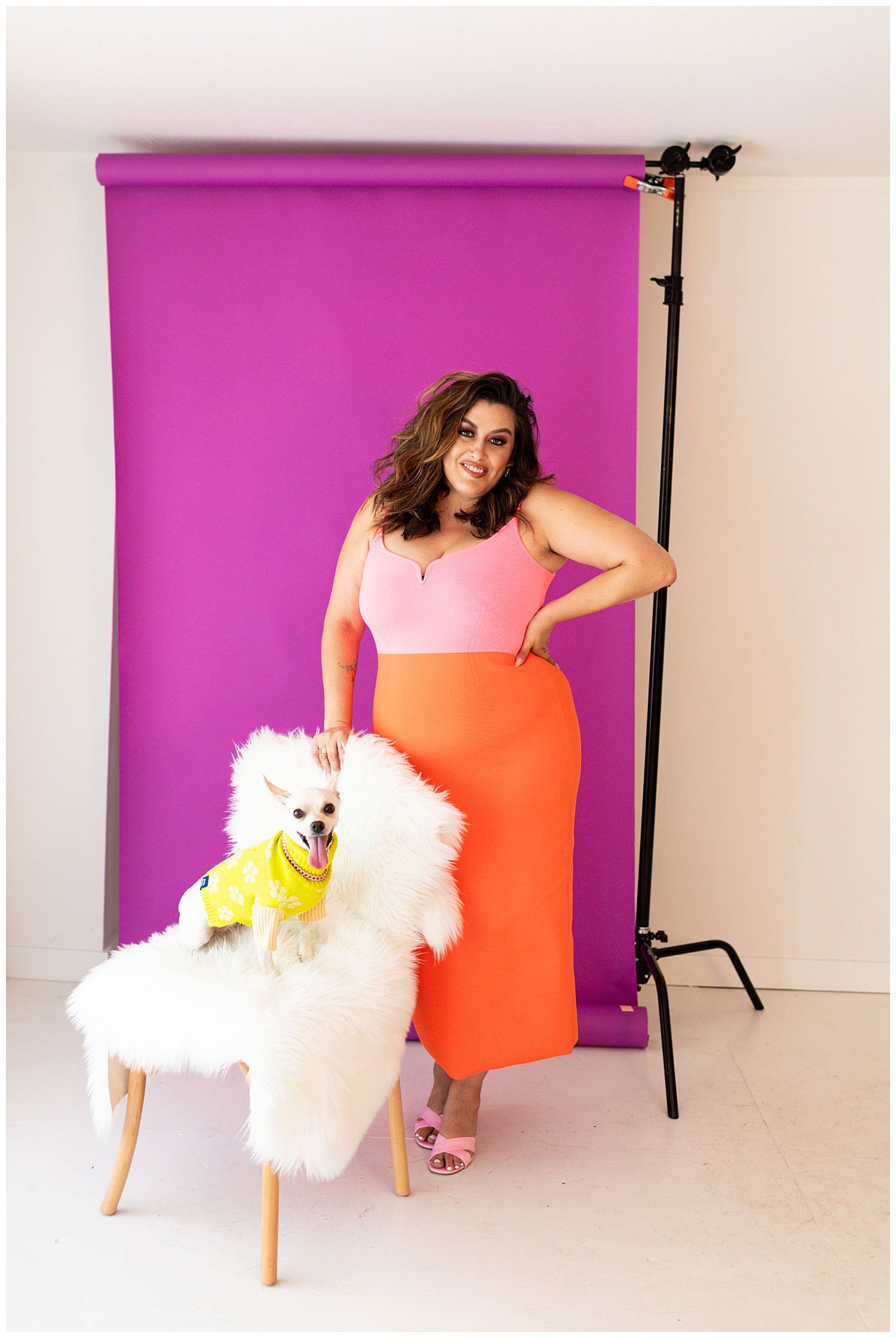 A woman in a pink and orange dress posing with a small dog on a furry white chair in front of a purple backdrop.