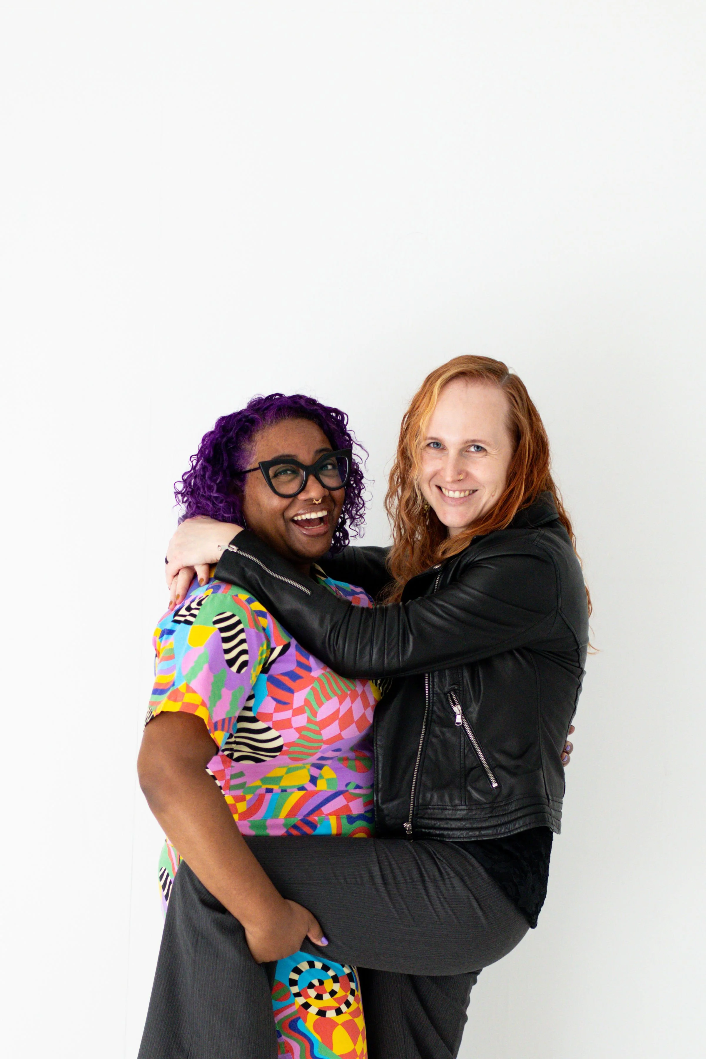 Two women embracing and smiling at the camera, one with purple curly hair and glasses, wearing a colorful patterned shirt, and the other with red hair, wearing a black leather jacket, against a plain white background.