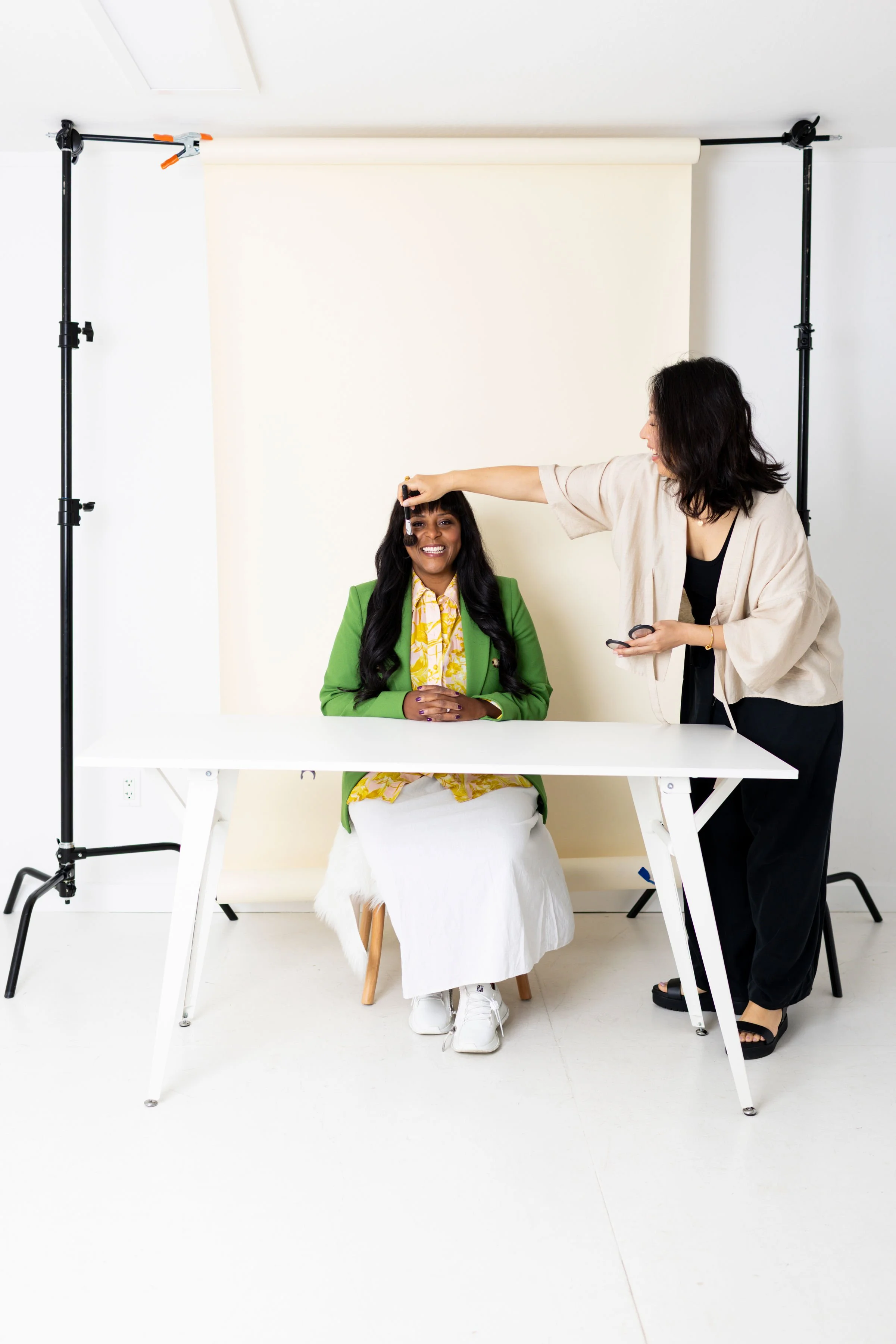 A woman sitting at a table getting her makeup done by another woman in a photography studio with a white backdrop and equipment.