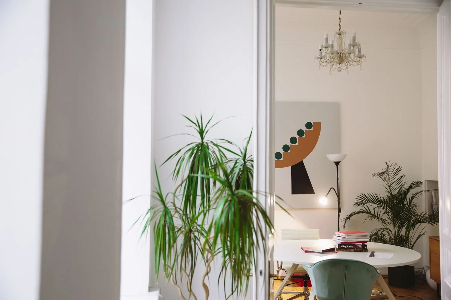 A bright interior dining area with a round white table, books, and an open laptop. There are large green potted plants, abstract artwork on the wall, a chandelier, and a standing lamp, all in a cozy, modern style.