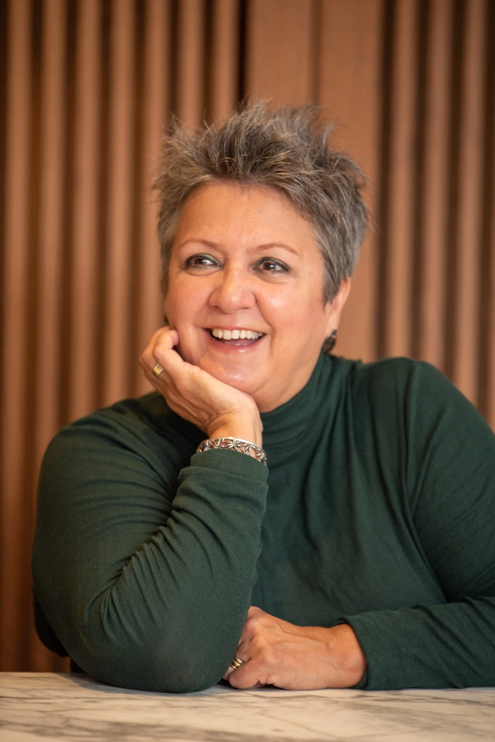 A woman with short gray hair smiling, sitting at a table with her hand on her chin, wearing a dark green long-sleeved shirt and a bracelet, with a wooden panel background.