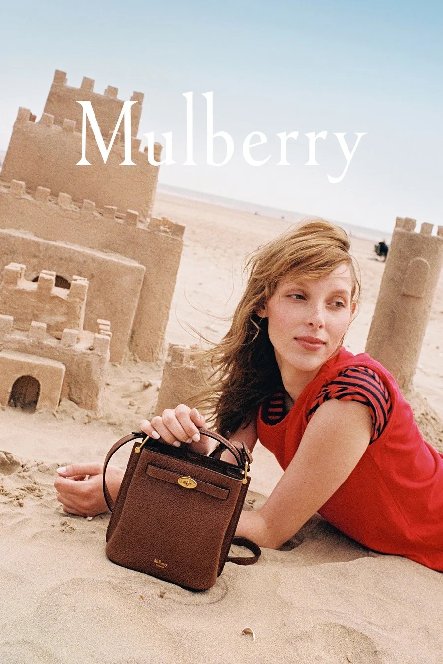 A woman lying on the sand at the beach, next to a sandcastle, holding a brown Mulberry handbag.