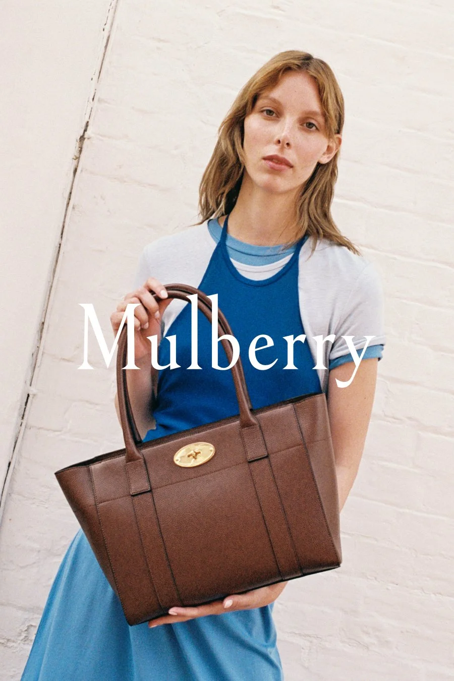 A young woman with shoulder-length light brown hair holding a brown handbag with a gold logo, standing against a white brick wall. She wears a blue dress layered with a white and blue shirt, and the text 'Mulberry' is overlaid on the image.