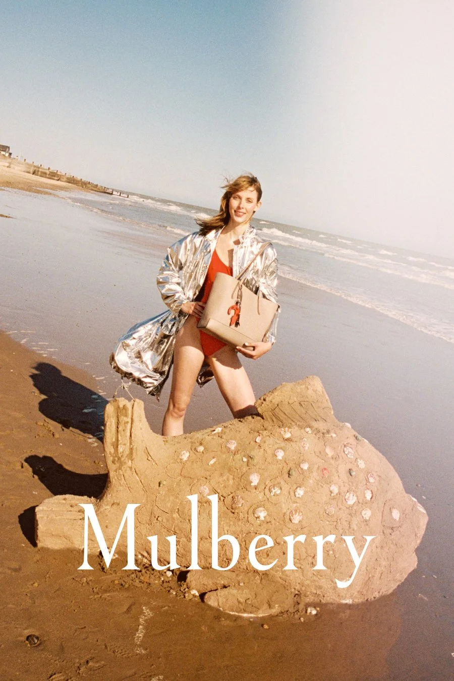 A woman standing next to a sand sculpture of a fish on a beach, holding a beige handbag with a red heart charm, wearing a shiny silver jacket and a red swimsuit, with the word 'Mulberry' written at the bottom.