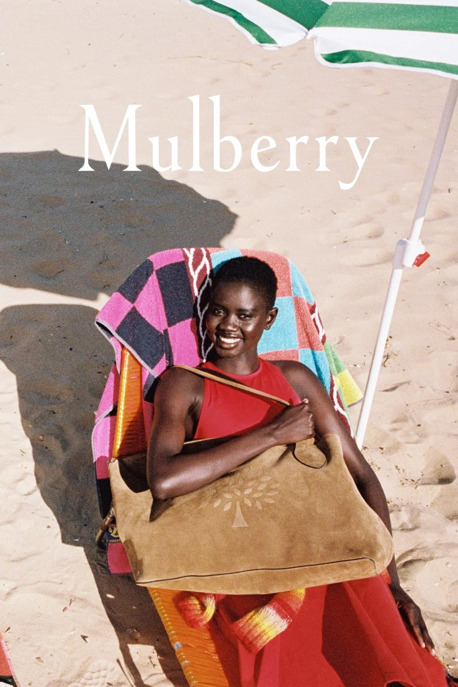 A woman sitting on a beach chair under a green and white beach umbrella, holding a large tote bag, with the word "Mulberry" overlayed at the top.