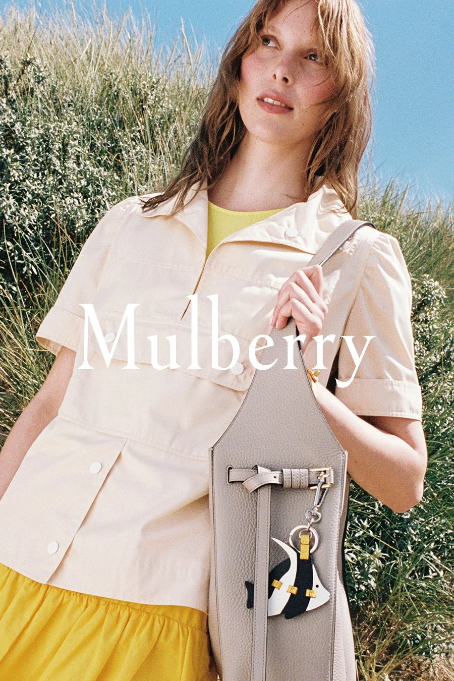 A woman outdoors with tall grass and a blue sky, wearing a cream-colored jacket, yellow dress, carrying a gray handbag with a Mulberry keychain.