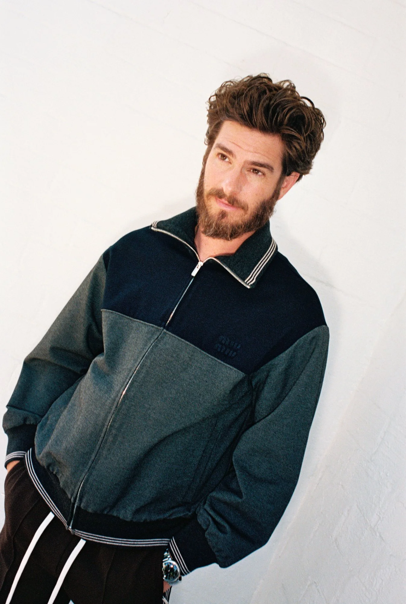 A man with brown hair, beard, and mustache, wearing a dark gray and navy fleece jacket, standing against a white textured wall, looking slightly to the left.