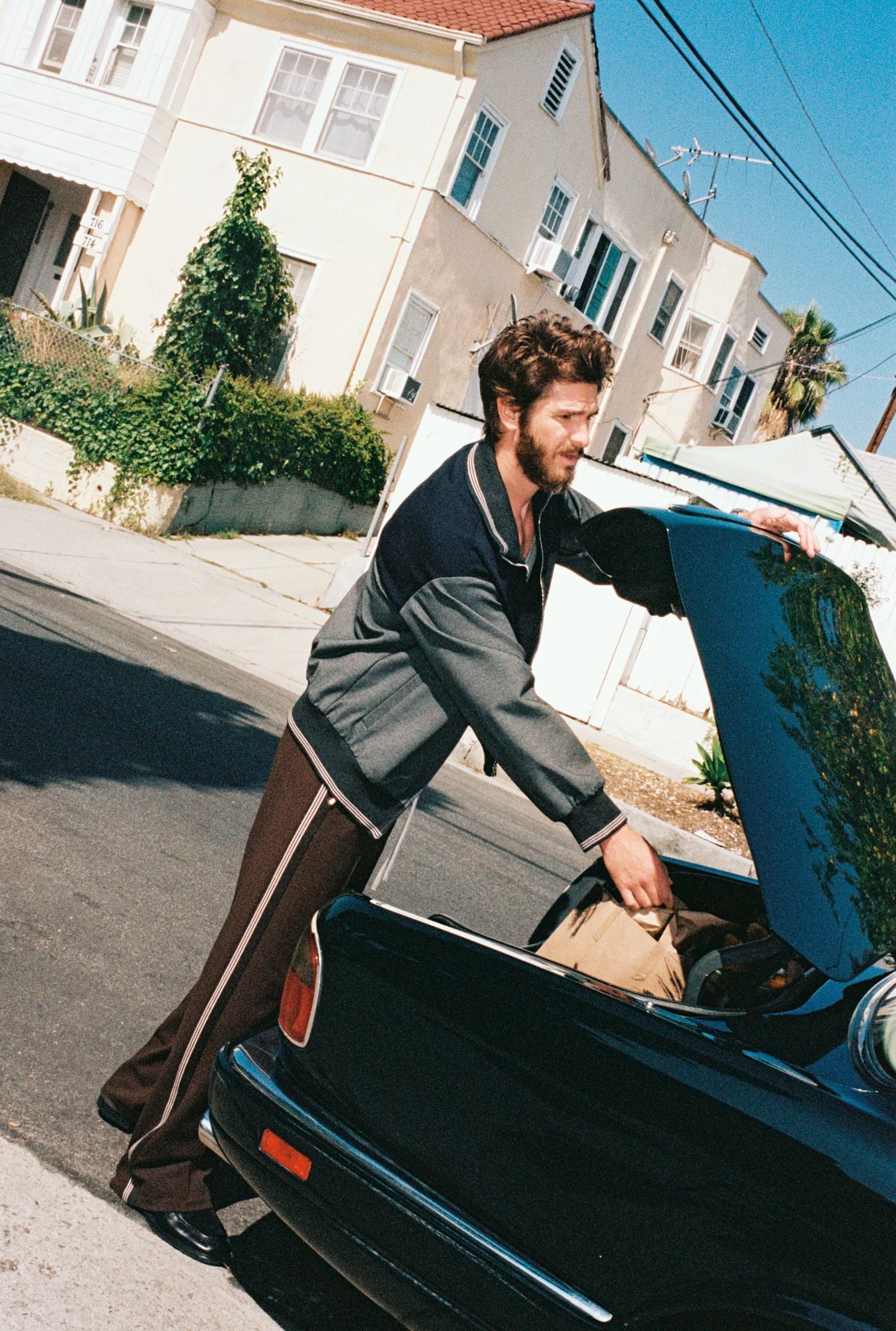 Man with brown hair and beard loading groceries into the trunk of a black car on a residential street with houses, trees, and power lines in the background.