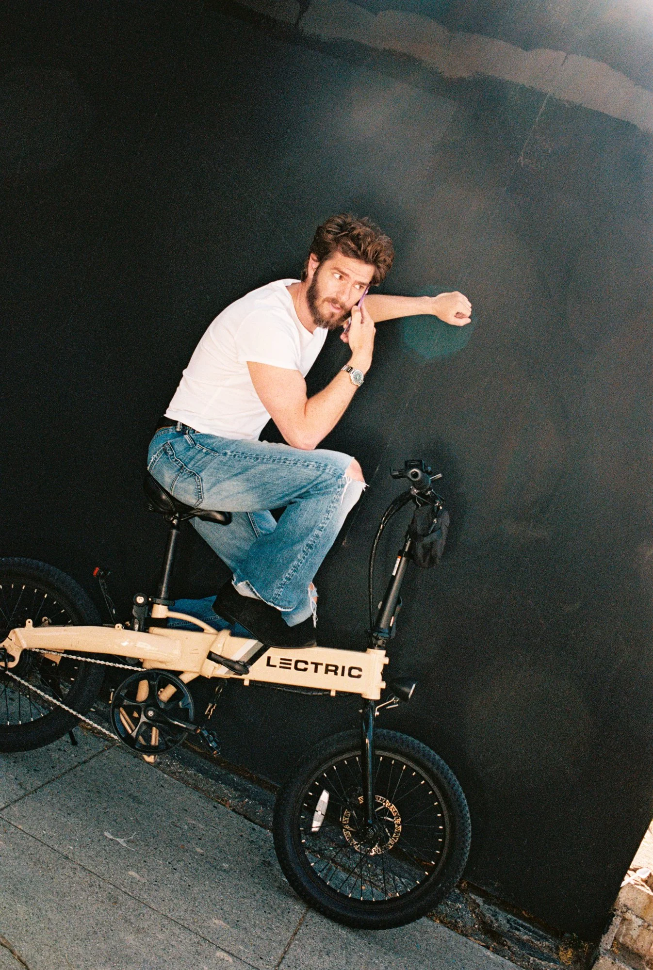 A man with brown hair and a beard riding a beige electric bicycle labeled 'LECTRIC' on a city sidewalk against a dark wall. He is wearing a white t-shirt, ripped jeans, and a watch, and appears to be talking on a cellphone.
