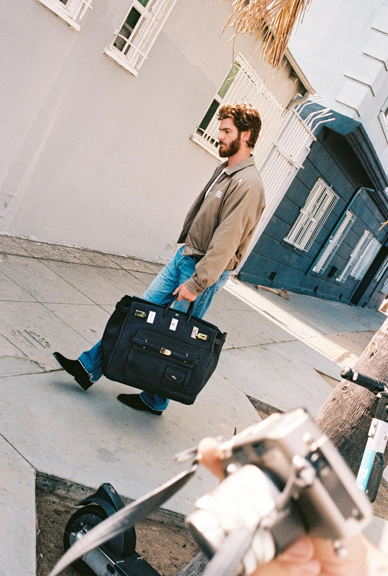 A young man with a beard walking on sidewalk, carrying a large black briefcase, wearing a beige jacket and blue jeans, with a lamppost and buildings in the background.