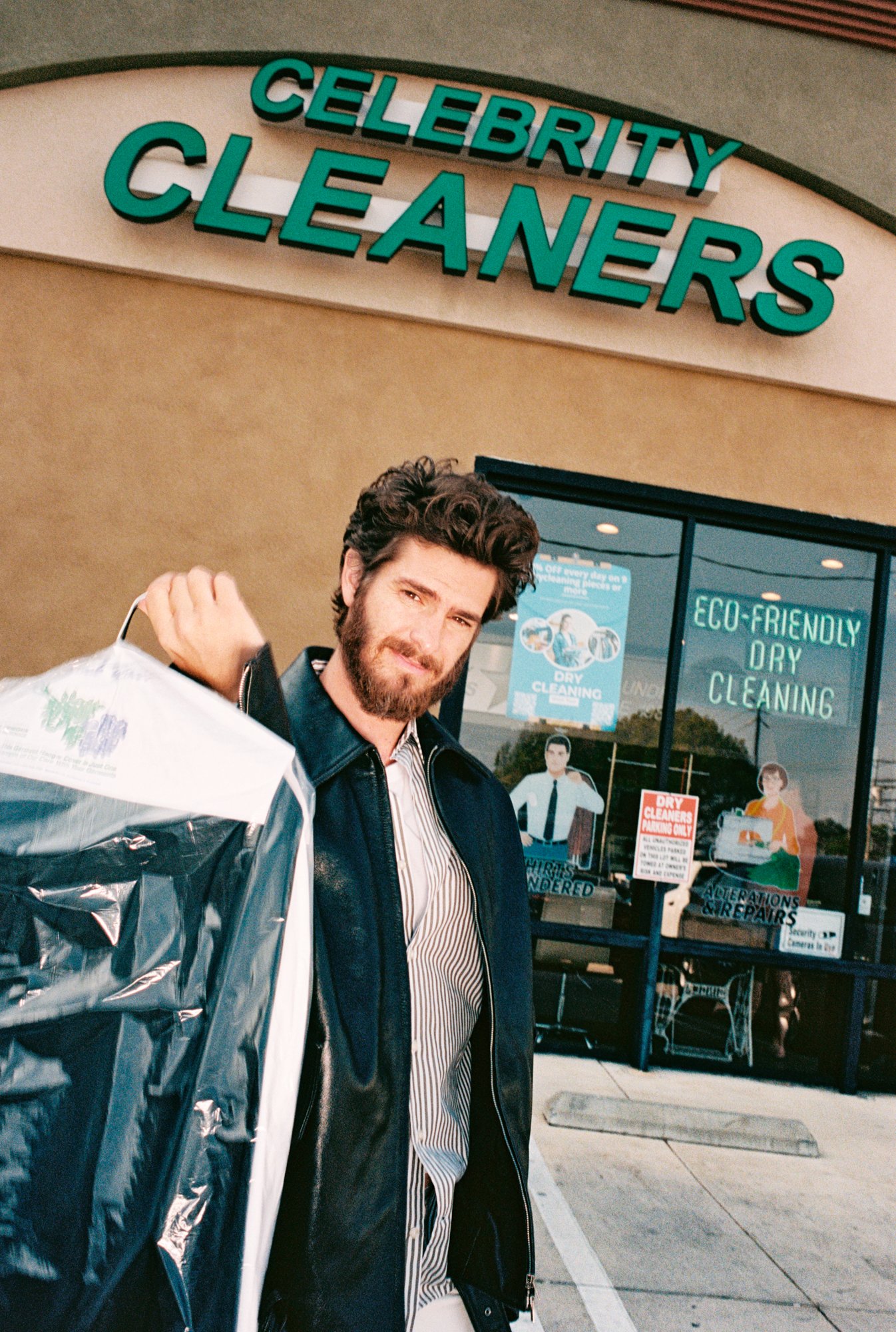 A man with dark curly hair and a beard standing outside a dry cleaning and alteration shop called 'Celebrity Cleaners.' He is holding a large plastic bag of laundry or dry cleaning and is looking at the camera. The shop has signs indicating eco-frien