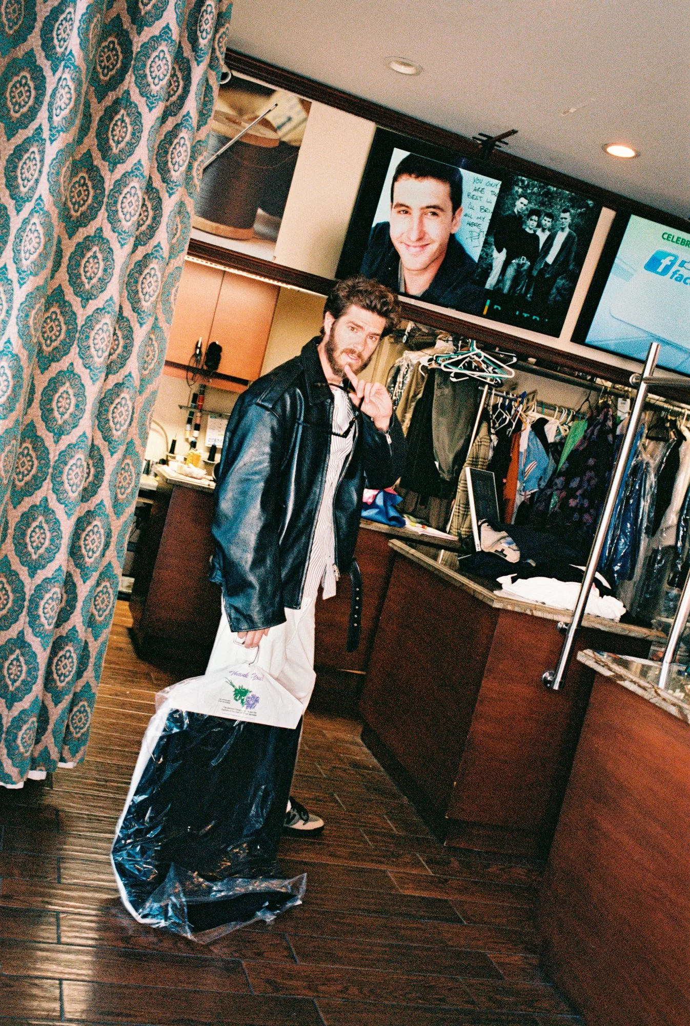 A young man with curly hair and a beard standing at a counter in a clothing store, holding shopping bags. There are two large screens on the wall behind him displaying images and text, and clothes hanging on racks in the background.