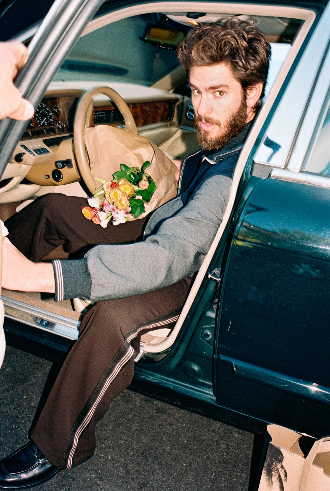A man with a beard and wavy hair sitting in the driver's seat of a vintage car, holding a bouquet of flowers wrapped in brown paper, with the car door open.