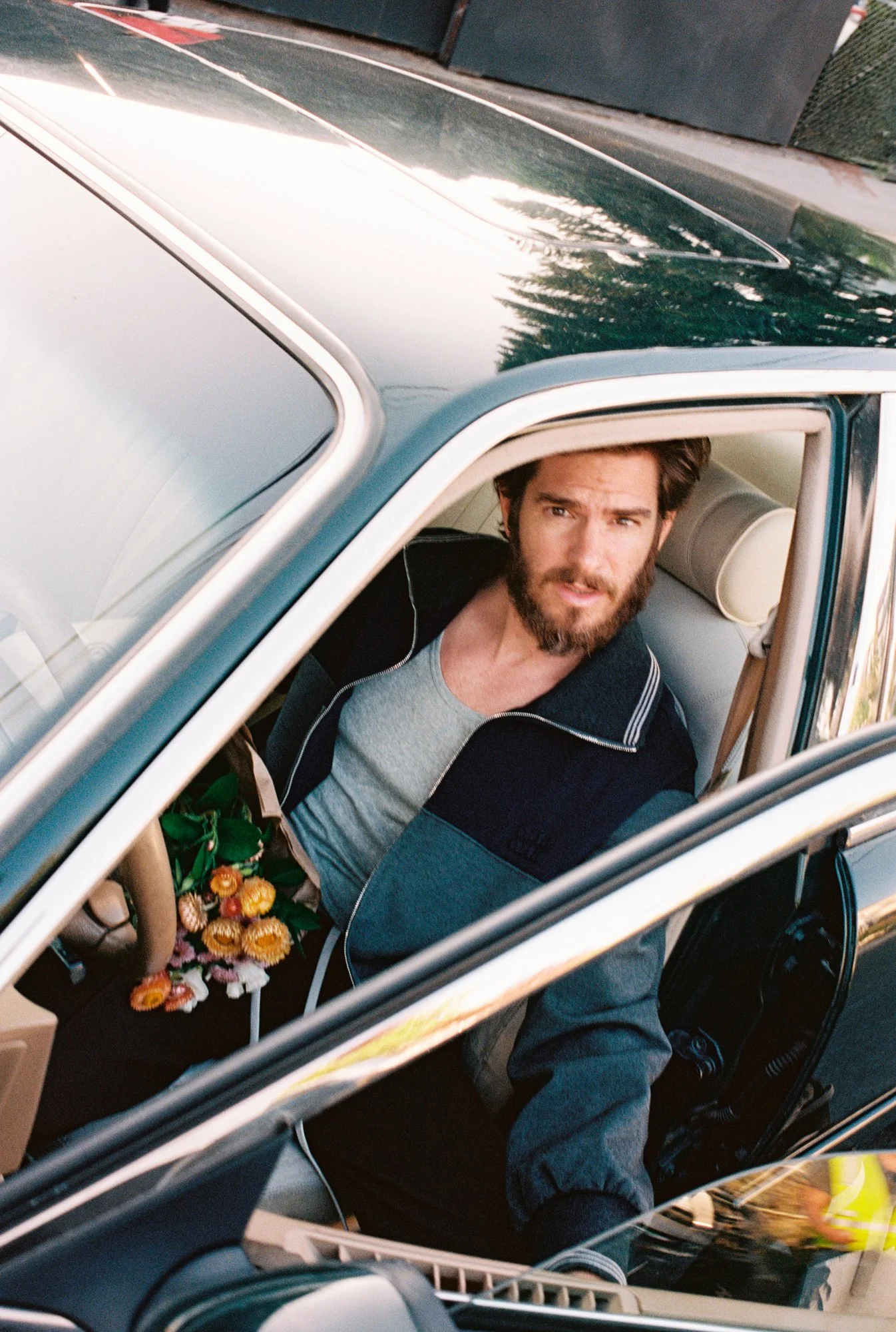 A man with a beard and brown hair sitting inside a car, looking out of the open window. The car seats are beige, and there is a bouquet of flowers on the dashboard.