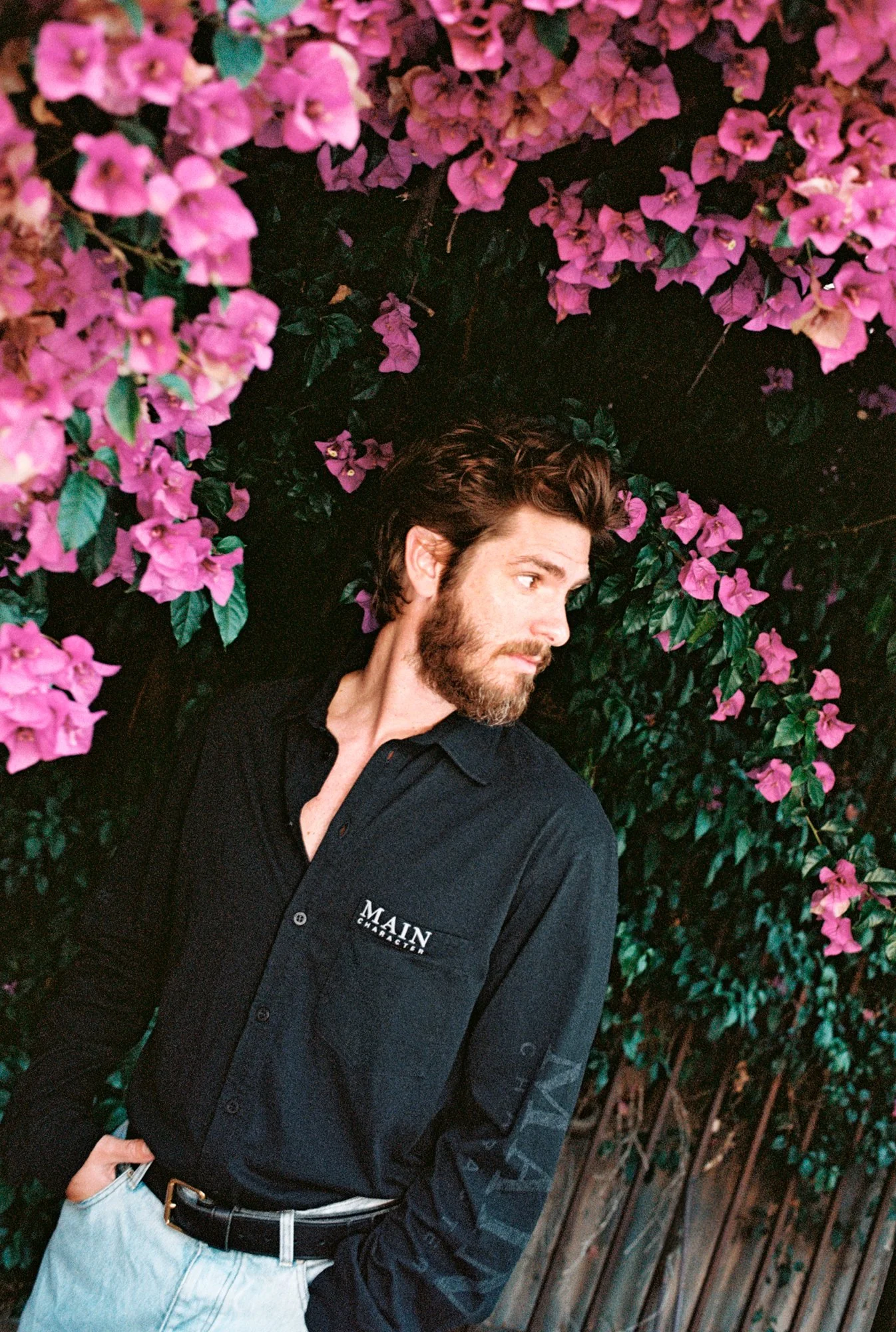 A man with brown hair and a beard wearing a dark blue shirt with 'MAIN CHARACTER' embroidered on it, standing next to a wooden fence covered with pink bougainvillea flowers.