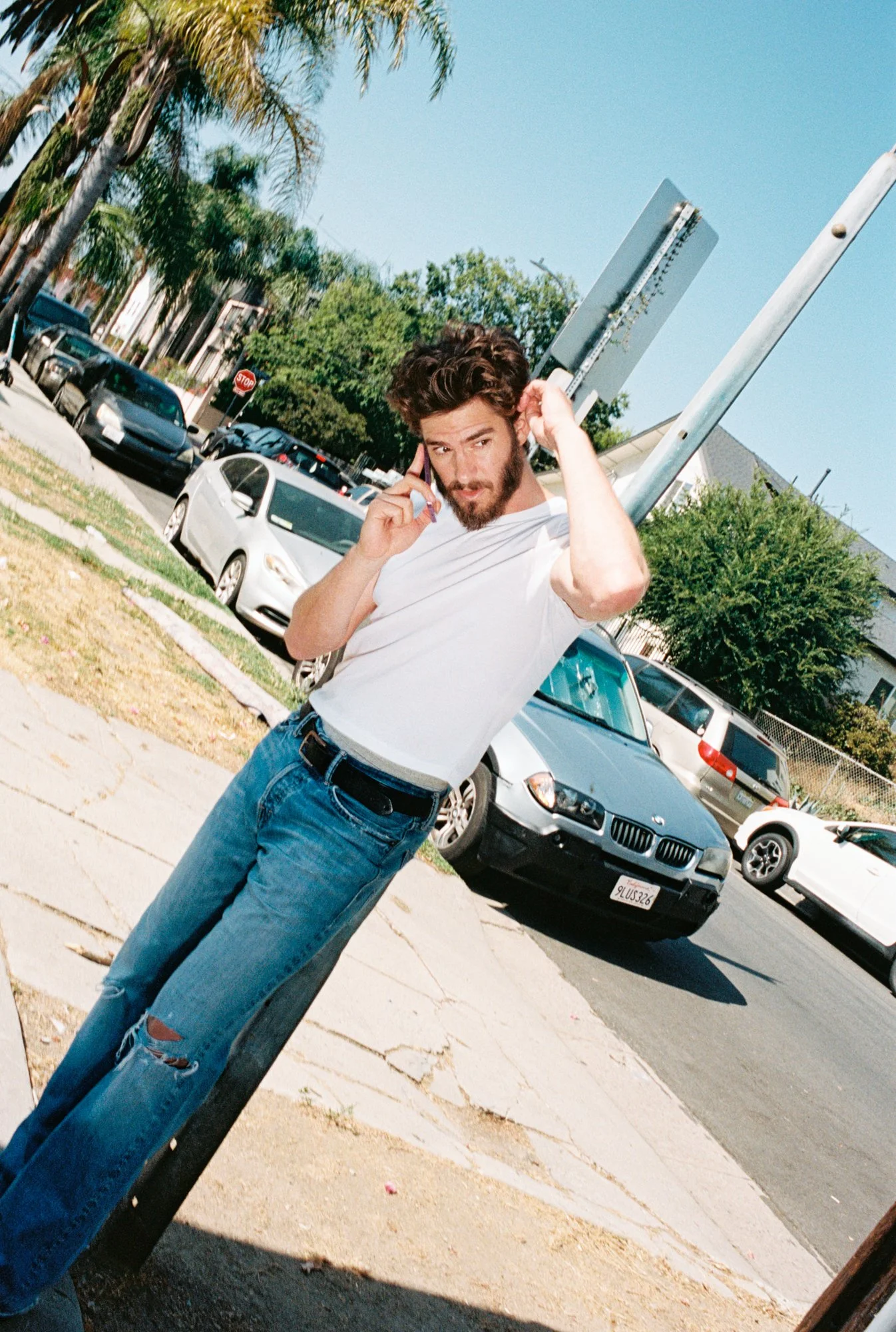A man with dark curly hair and a beard is talking on a cell phone while adjusting his hair with one hand. He is wearing a white t-shirt and blue ripped jeans. The background shows parked cars, palm trees, and a stop sign under a clear blue sky.
