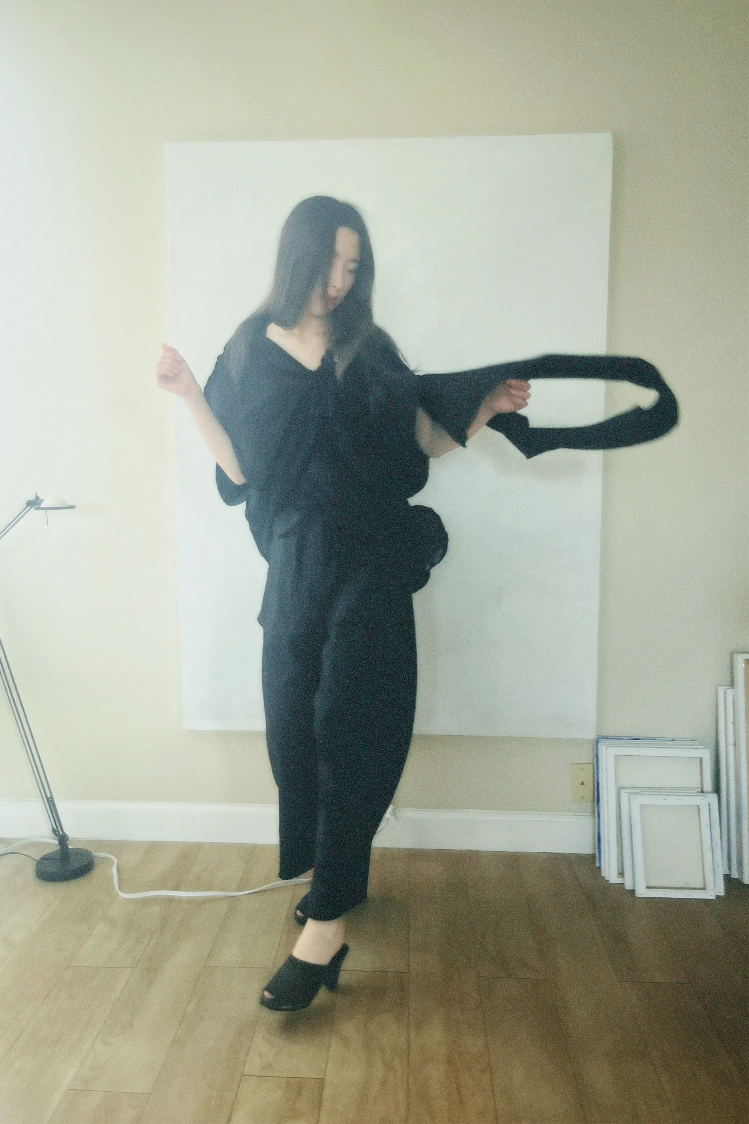 A woman with long black hair dressed in black clothing and high heels, standing on one foot with her other foot raised, holding a black leash or strap, in front of a white backdrop in a room with a wooden floor.