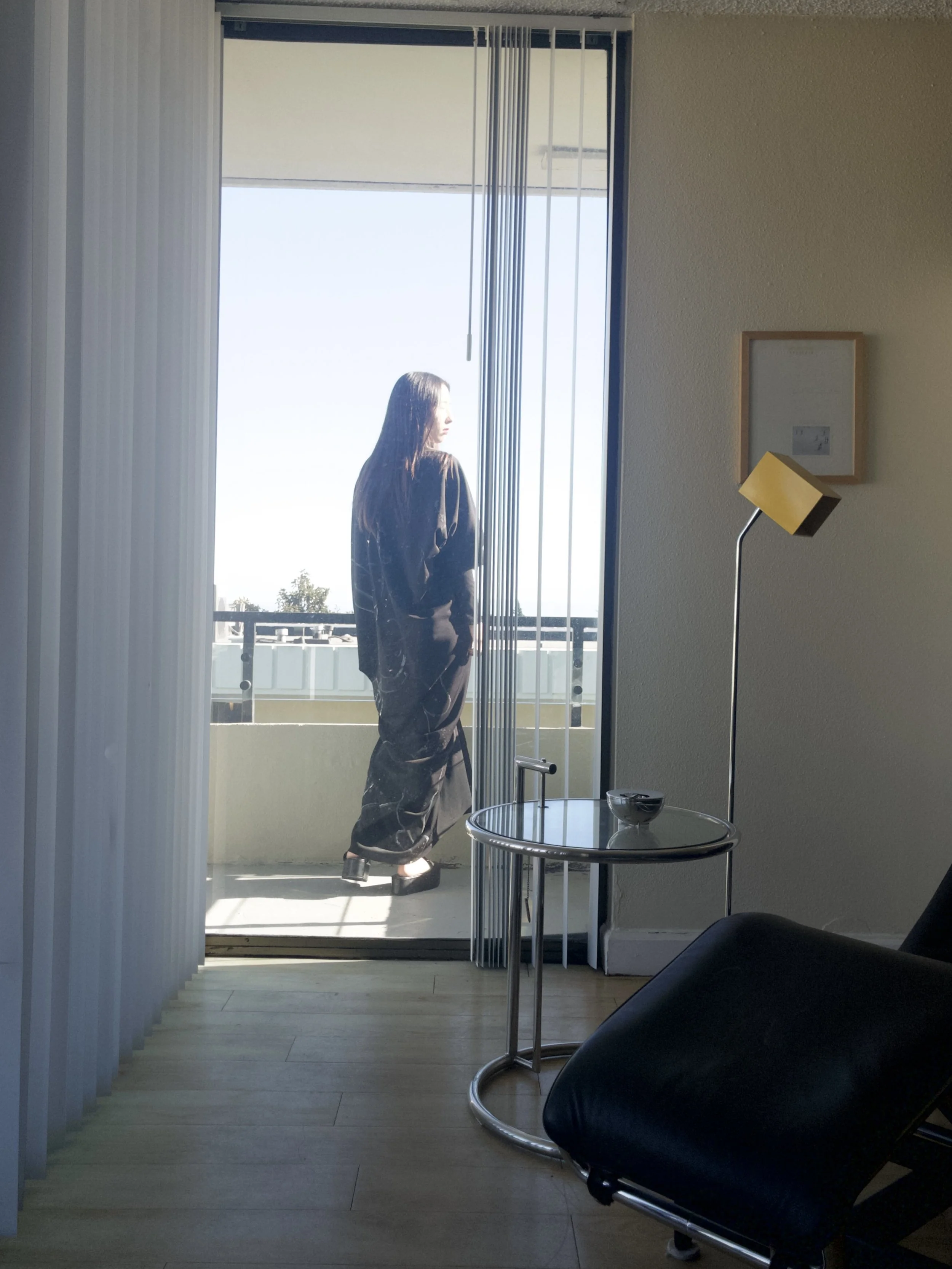 A woman in black pants and top stands on a balcony, looking outside. The room has a black leather chair, a small round glass table, a tall yellow lamp, and a picture frame on the wall.