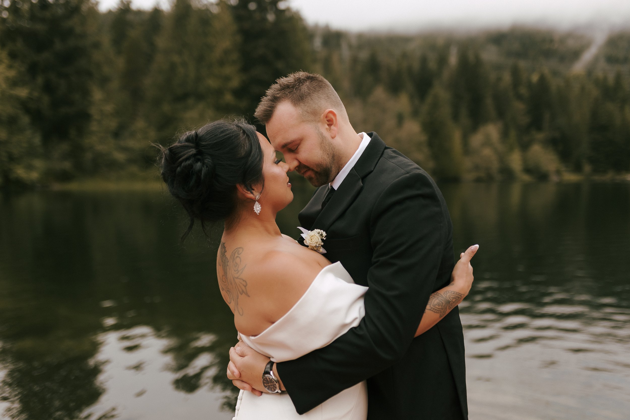 Couple embracing by a lake with trees in the background.