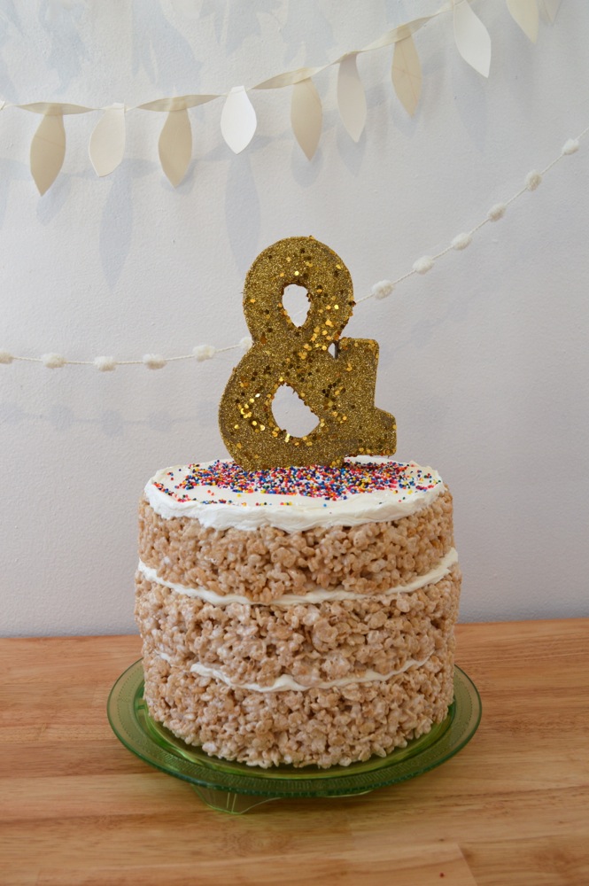 A naked cake made from layers of rice krispie treats, vanilla frosting, colorful sprinkles, and a glittery gold ampersand topper.