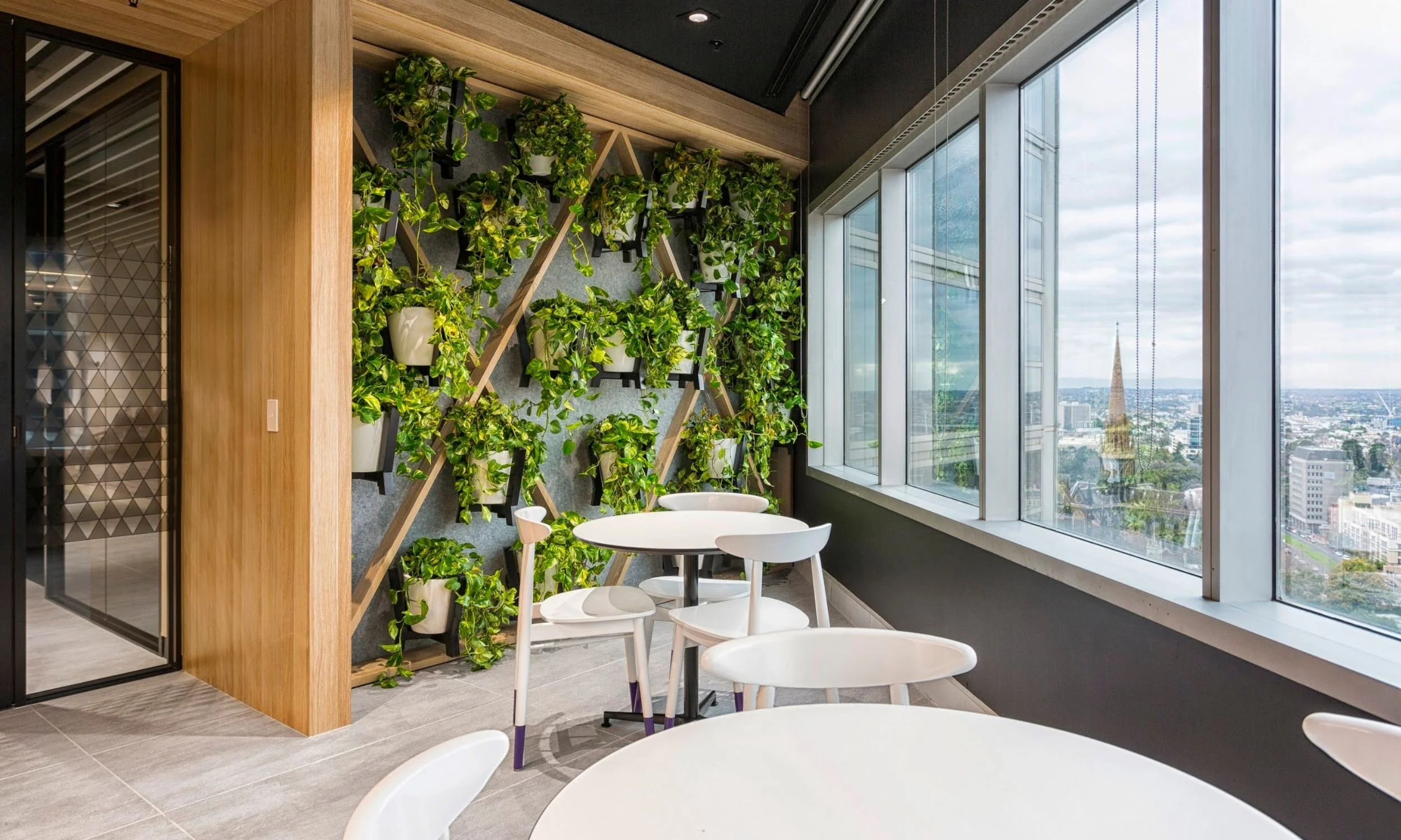 Modern Melbourne office breakout area featuring an indoor greenwall with cascading plants, white tables and chairs, and large windows overlooking the city skyline.
