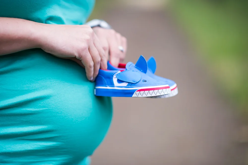Expectant mom holds tiny blue sneakers of her future baby next to her baby bump.