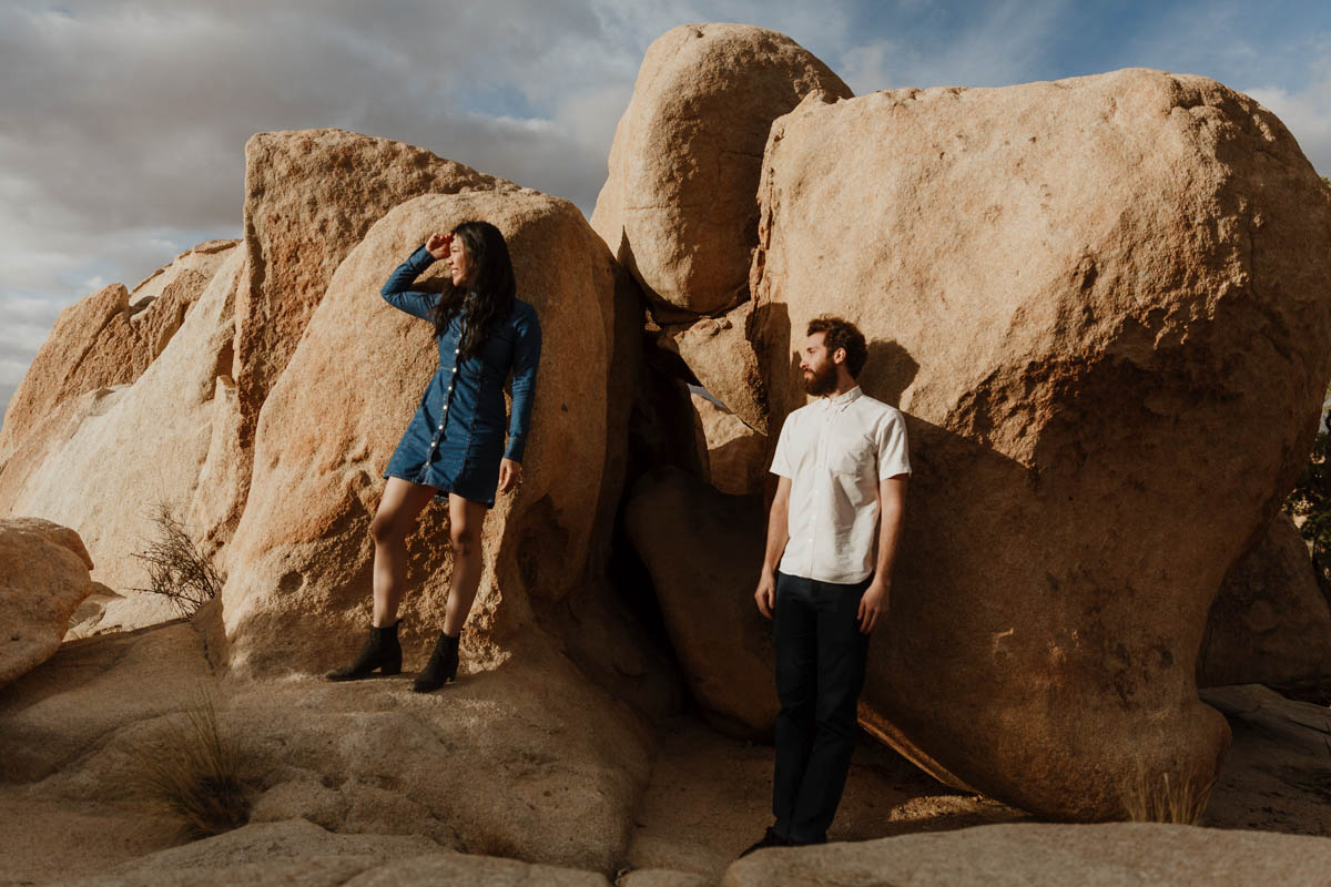 joshua tree california engagement session couple standing on rock cluster looking out at desert sunset