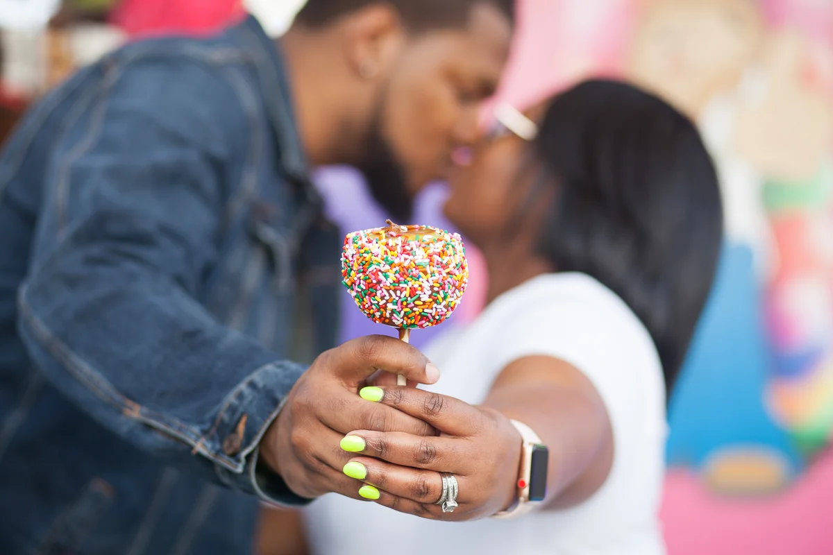 CARNIVAL PHOTO SESSION DURHAM NORTH CAROLINA BRITTANY AND DEON KISS WHILE HOLDING CANDY APPLE TO CAMERA