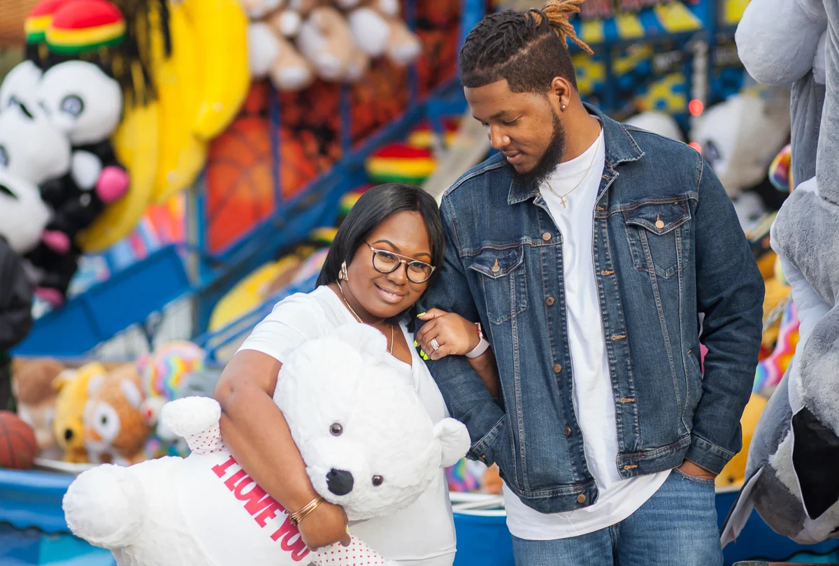 CARNIVAL PHOTO SESSION DURHAM NORTH CAROLINA COUPLE WALKING WITH ARMS LOOPED, BRITTANY HOLDING STUFFED BEAR WEARING A T-SHIRT THAT READS "I LOVE YOU"