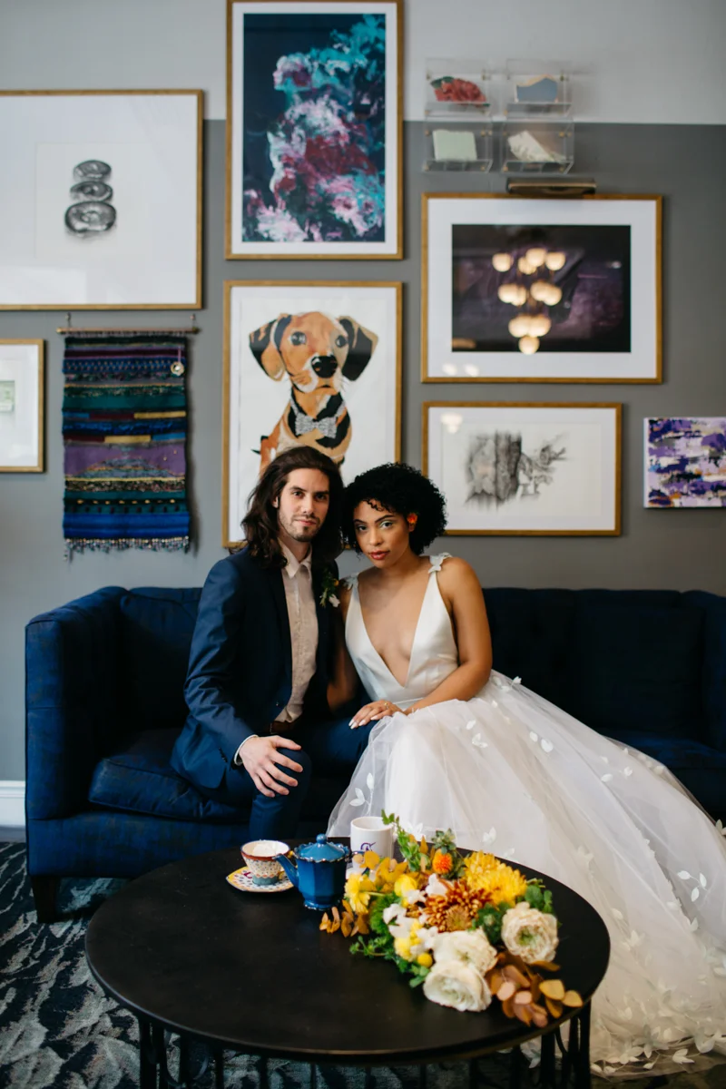 wes anderson inspired wedding styled shoot columbia south carolina bride and groom on sofa, bouquet and mismatched tea set on table, portraits on wall behind them