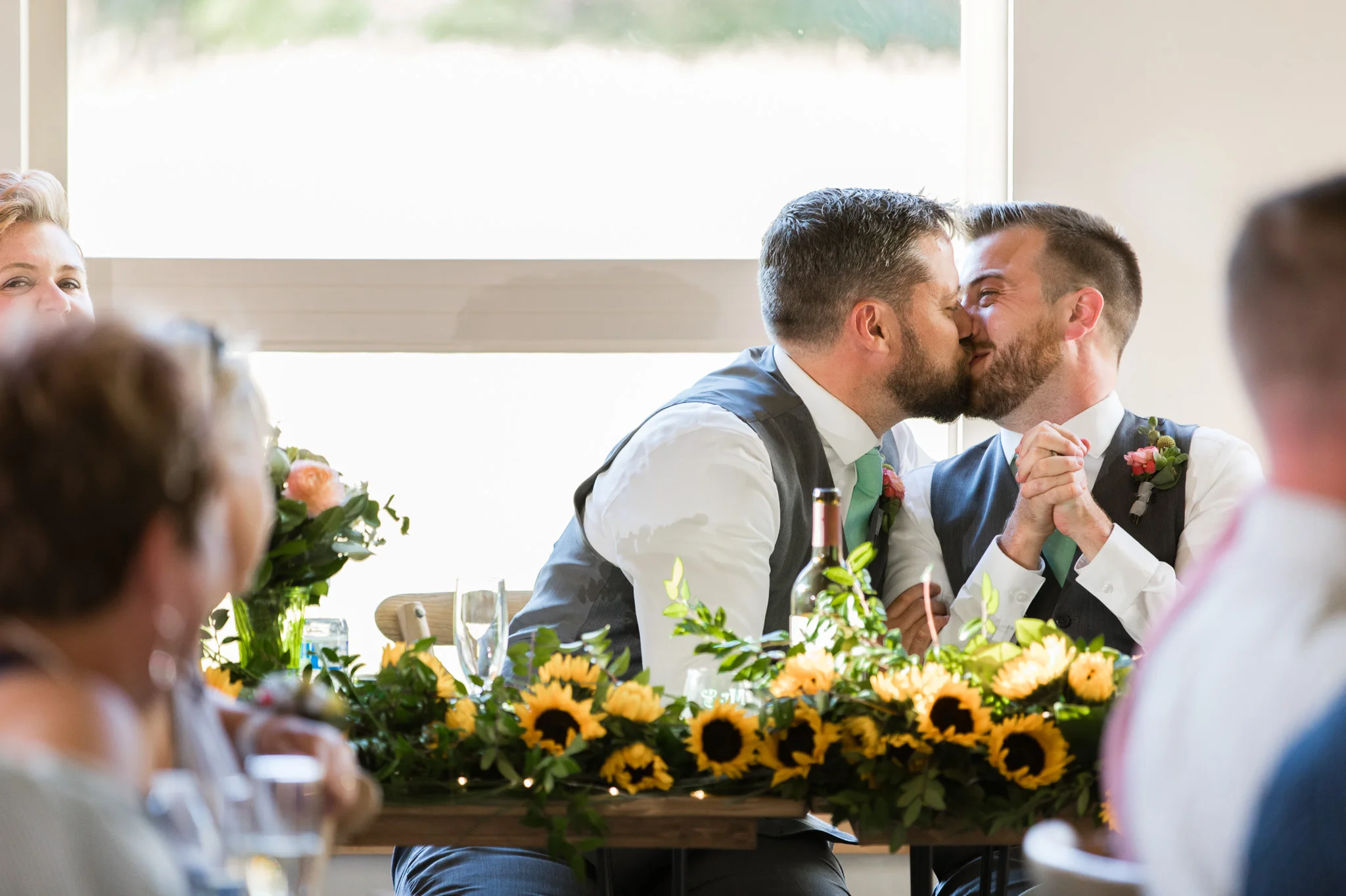 two grooms sharing a kiss at the head table during their wedding reception with sunflowers on the table Meera Graham Photography Missoula Montana