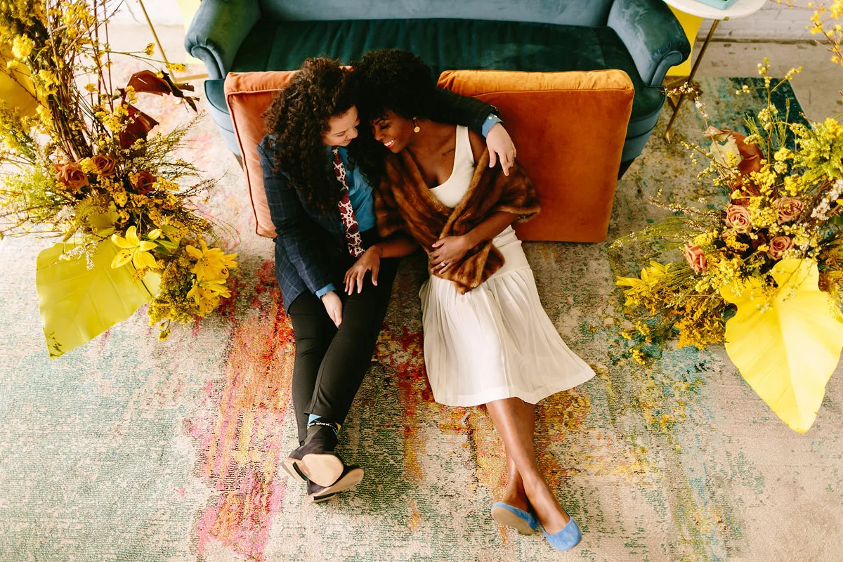 wes anderson inspired wedding brooklyn new york couple sitting on floor against couch with flowers on either side
