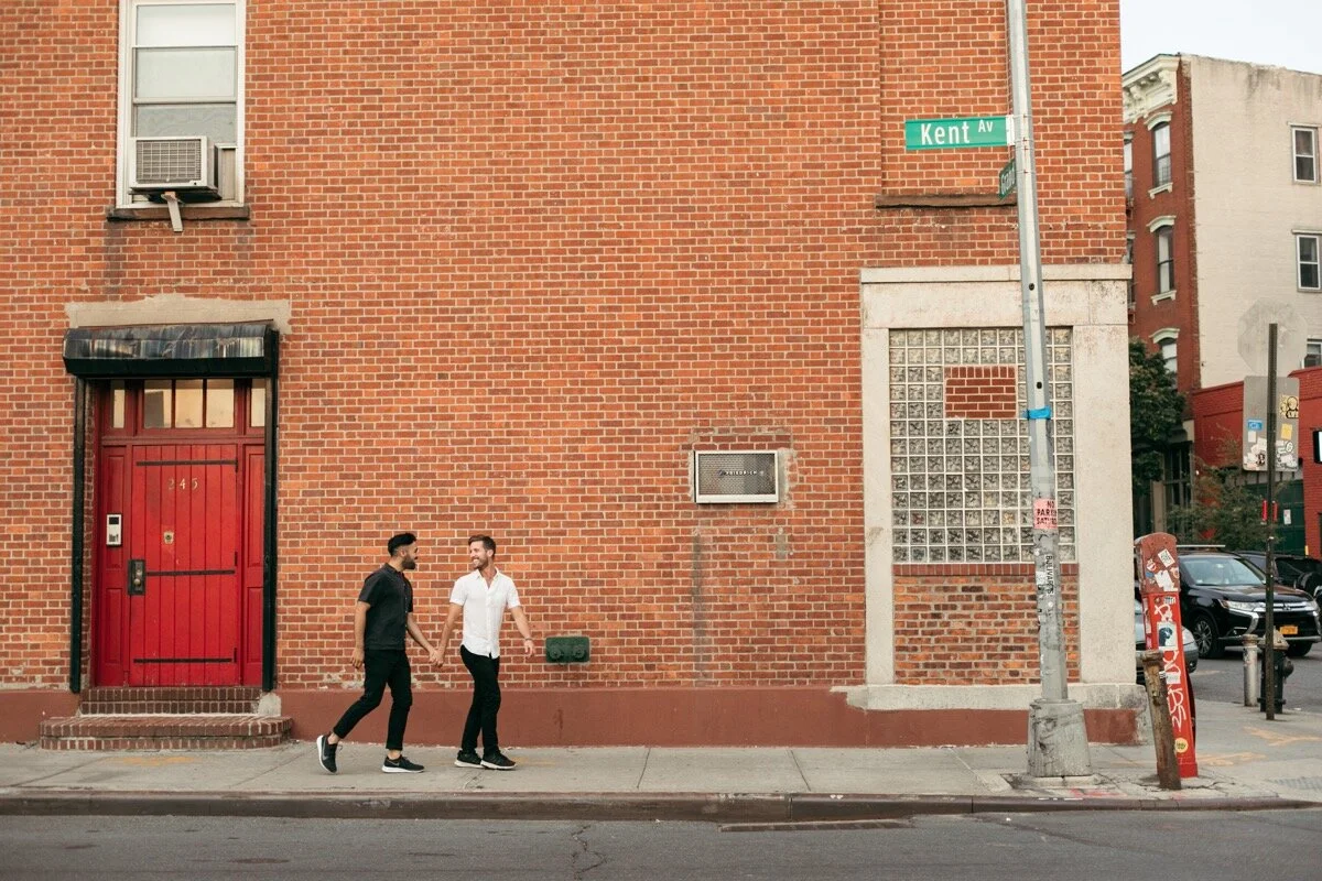 Joe and Rohan walk down the sidewalk holding hands during couples photo session in Domino Park Brooklyn Bailey Q Photo