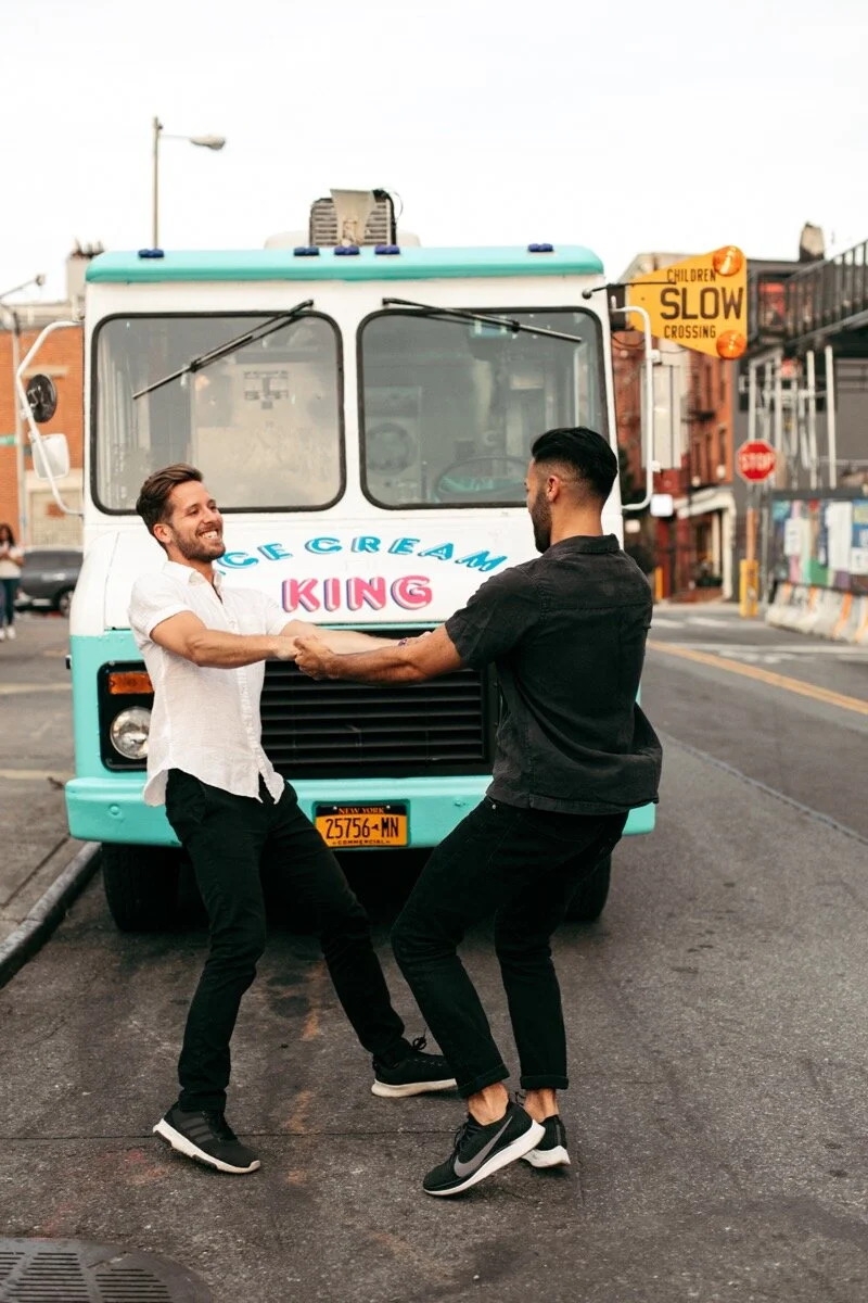 Joe and Rohan twirl around holding hands in front of ice cream truck during couples photo session in Domino Park Brooklyn Bailey Q Photo